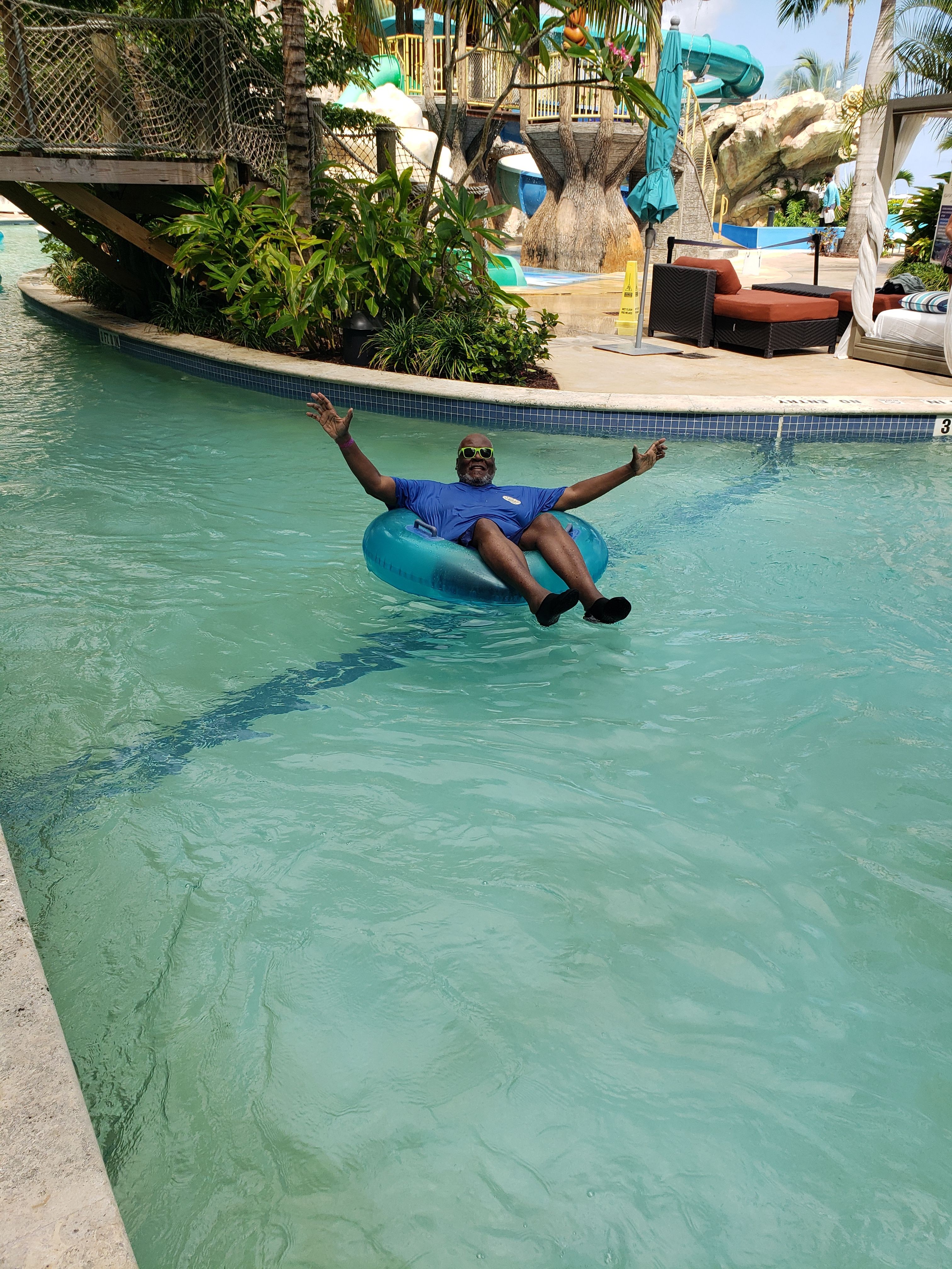 Child in blue swimsuit floating on a pool float, reaching towards the pool edge, surrounded by greenery and poolside furniture.