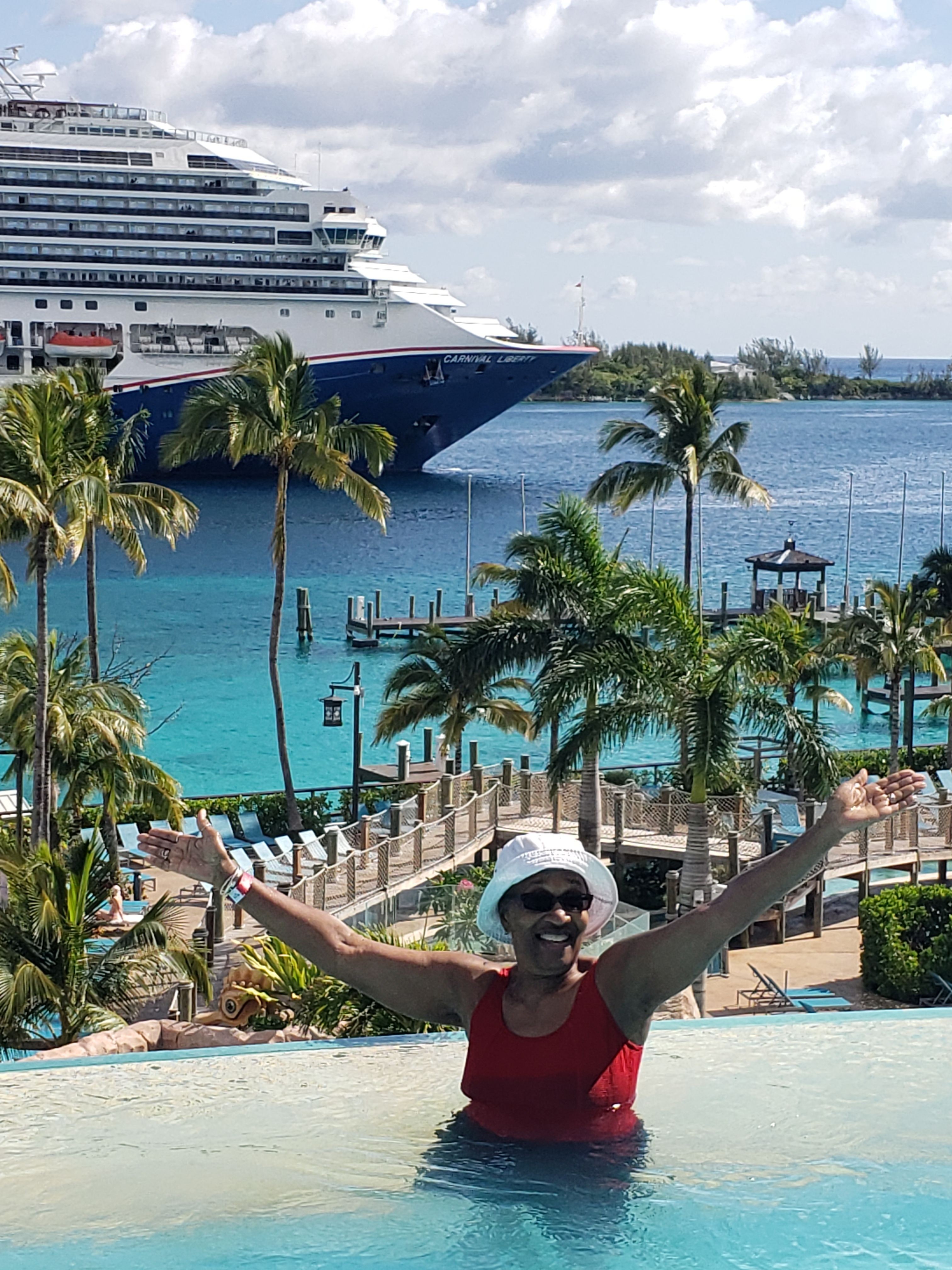Person in a pool wearing sunglasses and a hat, with a tropical resort background including palm trees and a large boat.