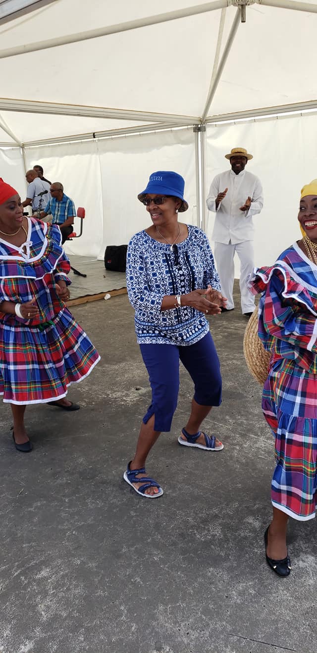 People dancing in traditional colorful clothing under a tent, with a woman in a blue hat and patterned top, and others in traditional attire, smiling and enjoying the event.