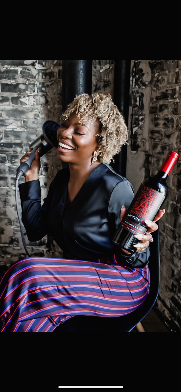 Woman with curly hair smiling, holding a wine bottle and a microphone, sitting against a brick wall.