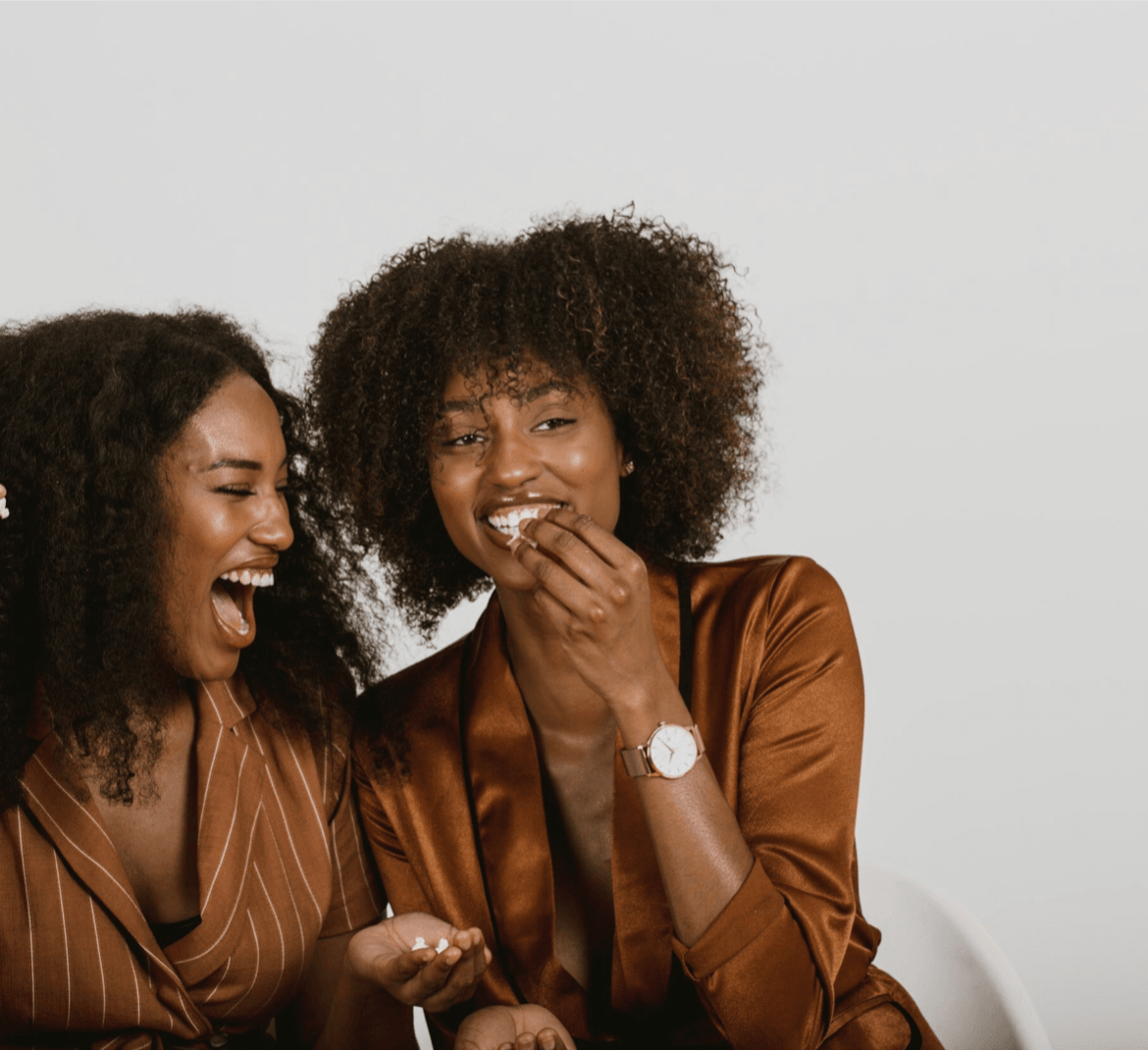Two women laughing and smiling, one covering her mouth, against a plain light background.