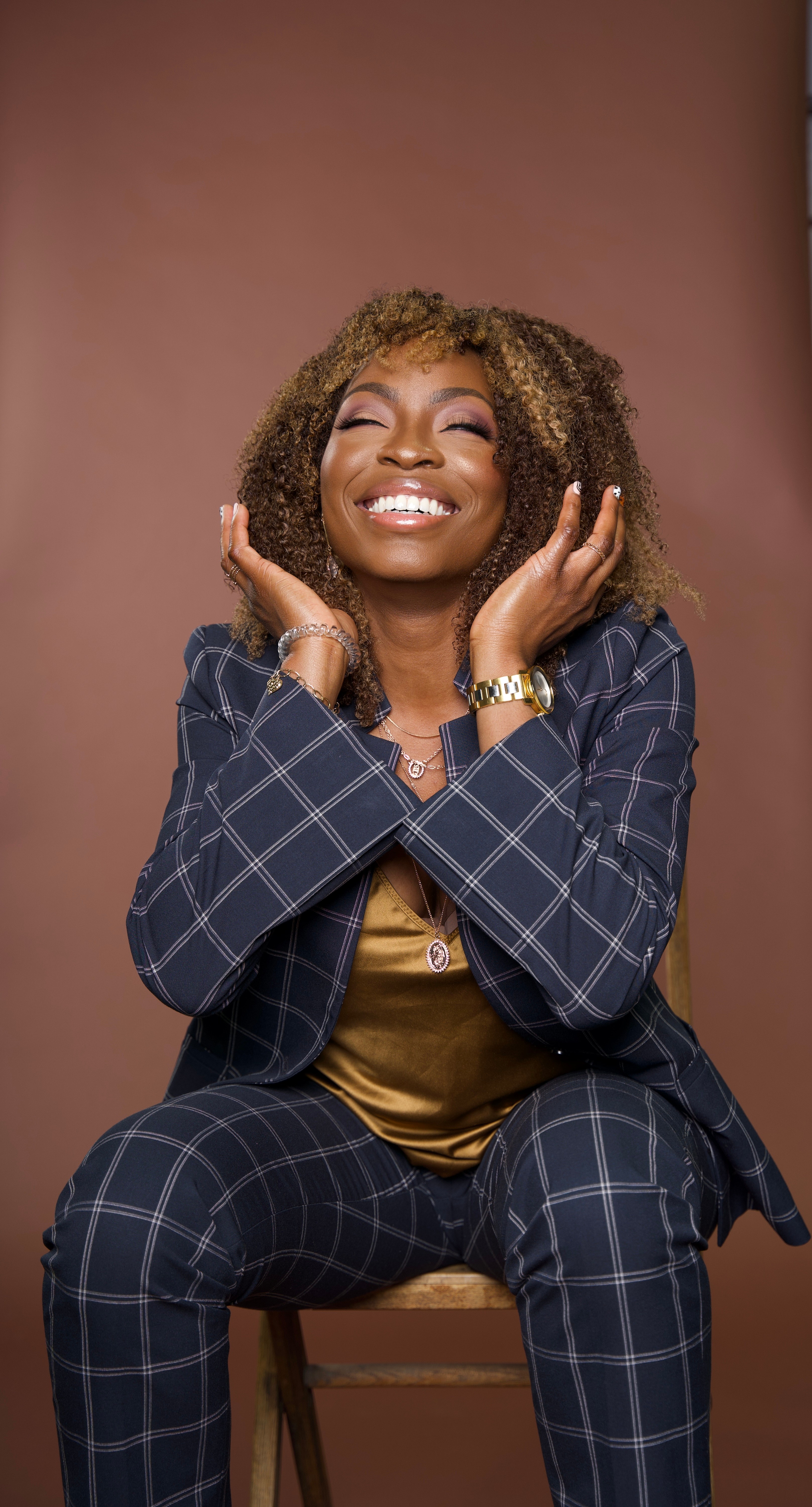 Smiling woman with curly hair, wearing a checkered suit, sitting on a chair with hands near her face.