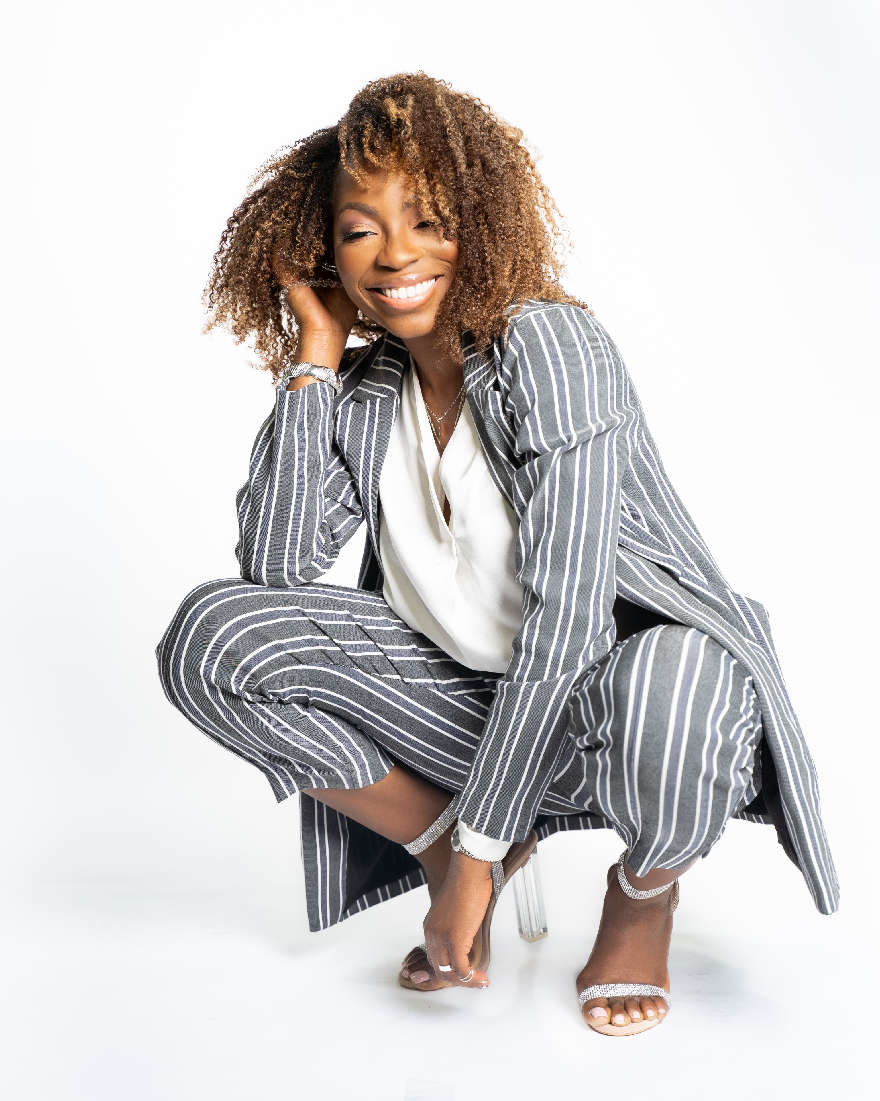 Woman crouching, smiling, wearing a striped suit and white blouse, with curly hair, against a white background.