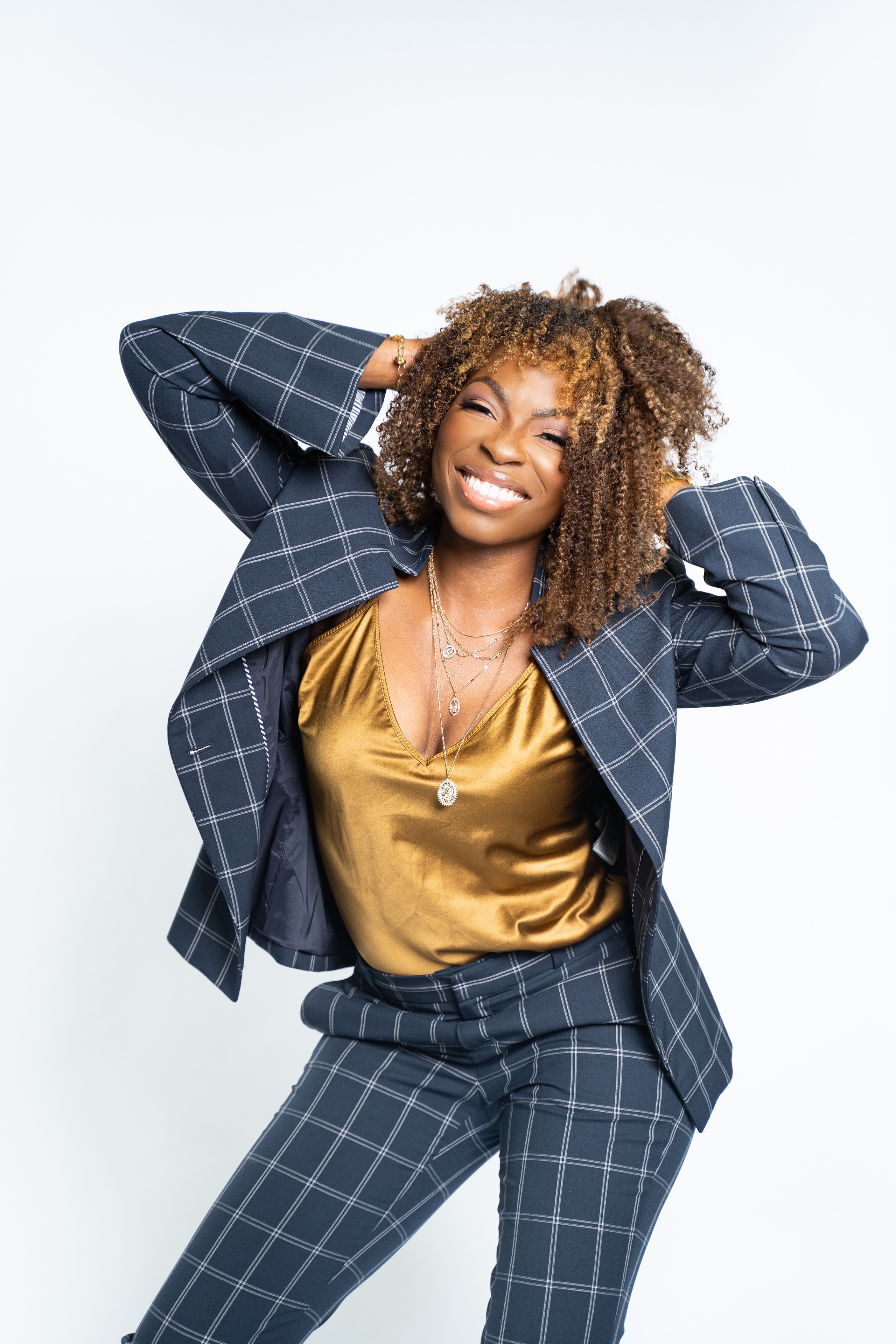 Smiling woman with curly hair wearing a checkered suit and gold top, posing with hands behind head against a white background.