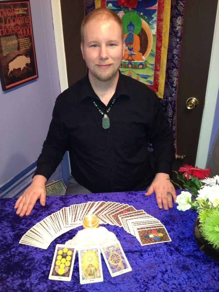 Young man with short hair and a black shirt stands behind a table with tarot cards and a crystal ball, smiling.