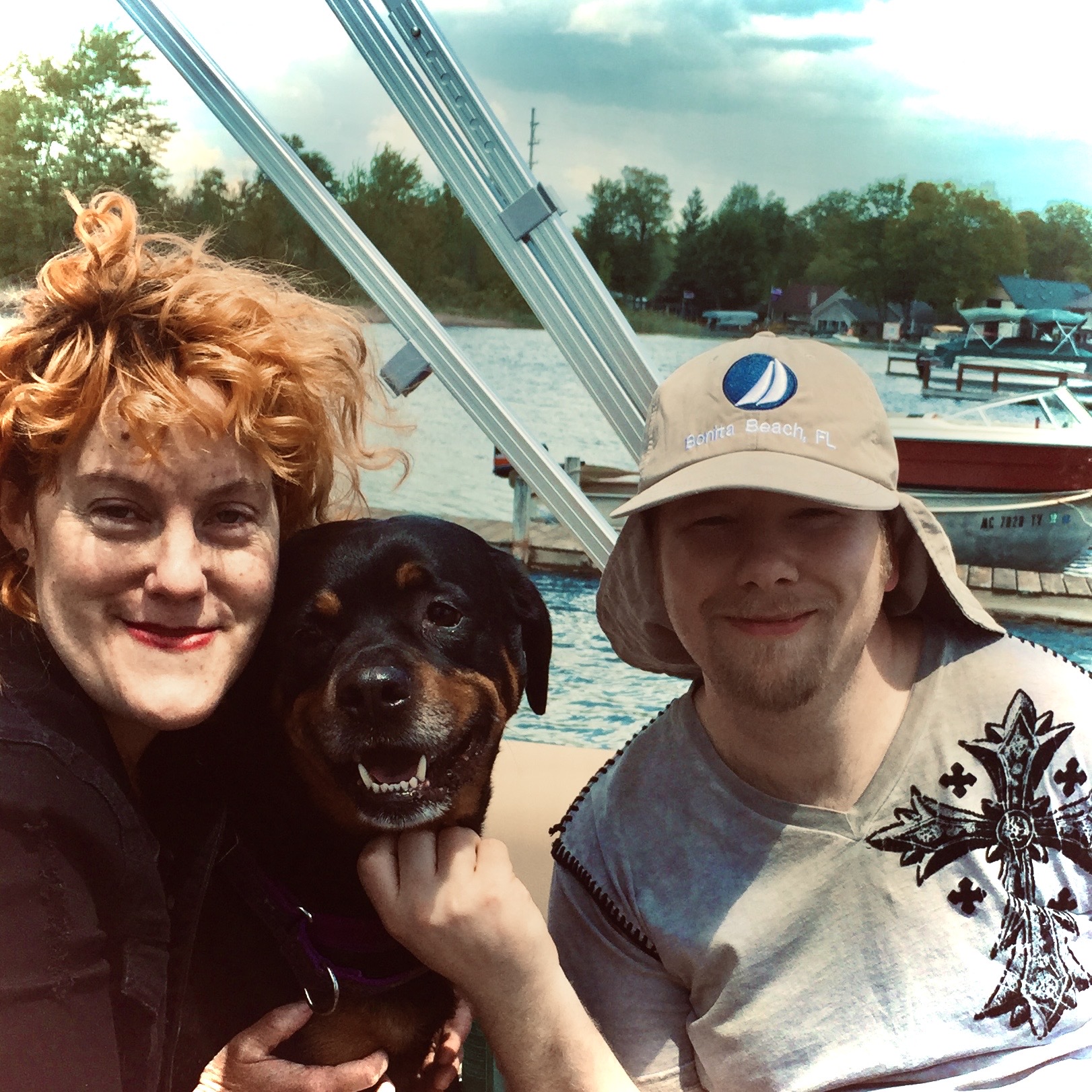 Two people and a dog at a marina with boats and water in the background.