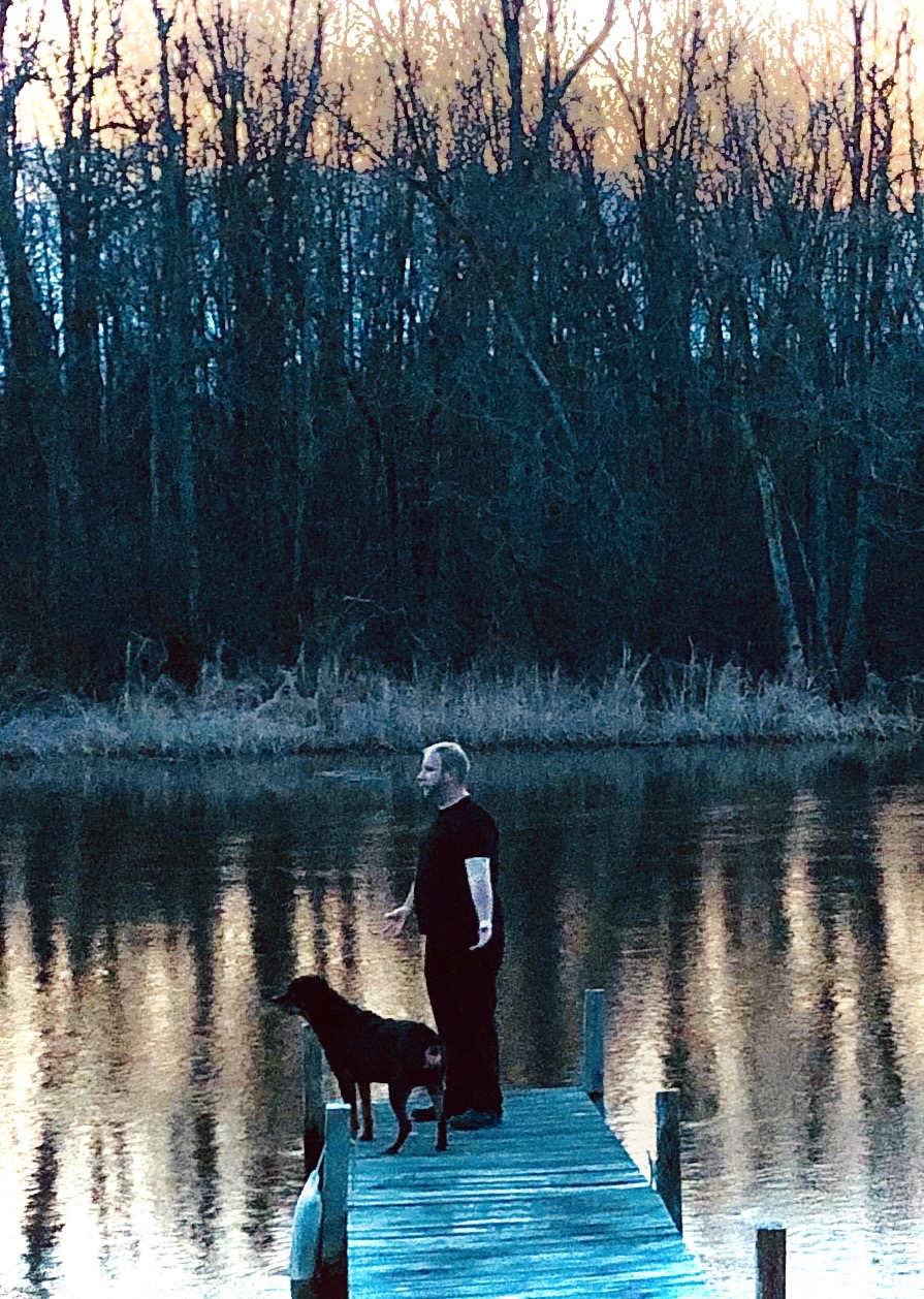 Person standing on a dock with a dog by a calm river, trees in the background, during sunset or sunrise.