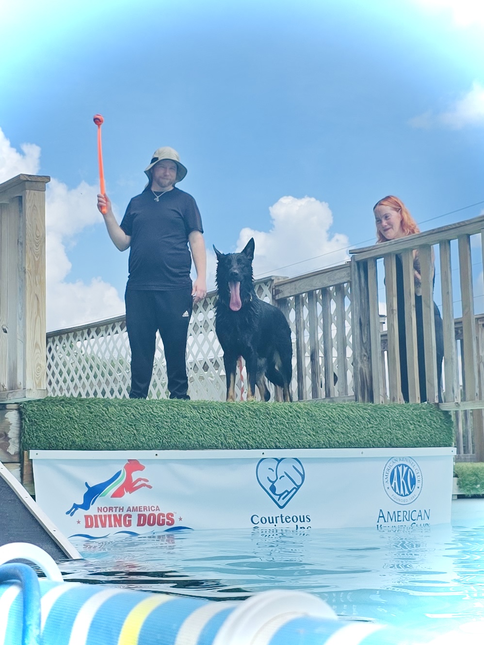 Person with a hat holding a paddle, a dog, and a woman on a deck overlooking a pool, with a blue sky and clouds.