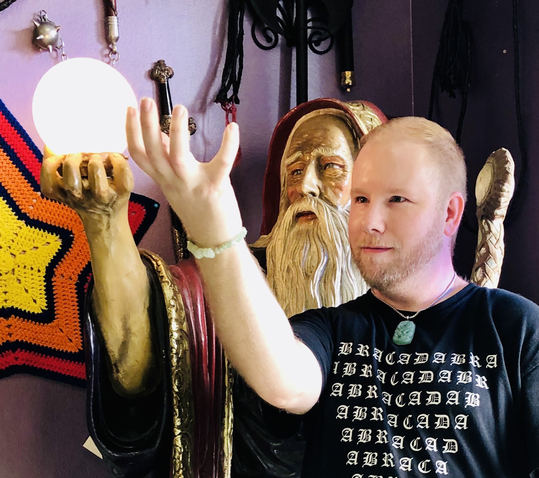 A man with a beard and a black shirt stands next to a statue of a bearded figure holding a mirror, touching the mirror.