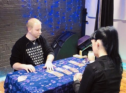 Two women sit at a table with a blue tablecloth, playing a card game against a dark brick wall with blue string lights.
