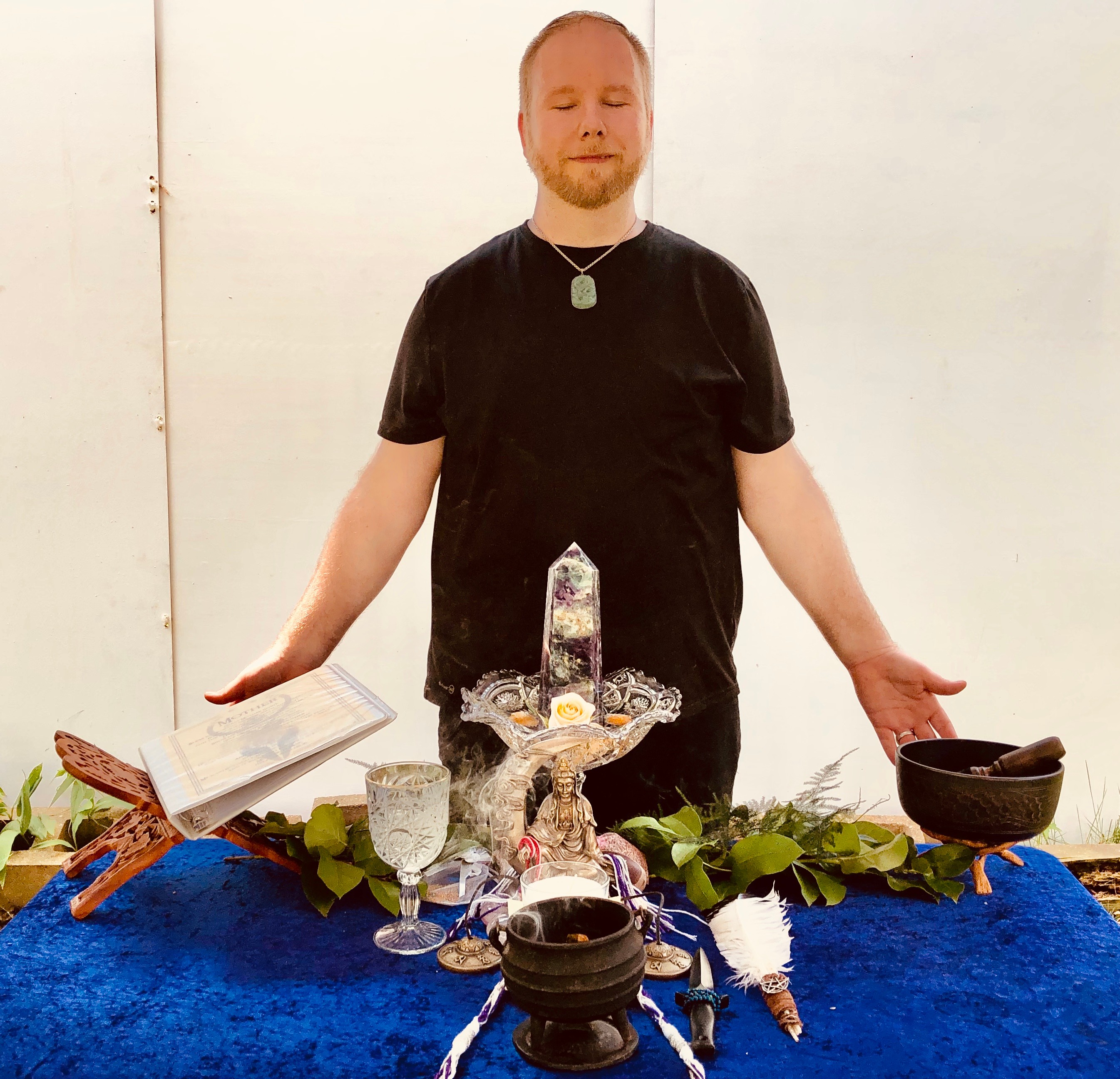 Person with eyes closed standing behind a table with various objects, including a crystal, glasses, and a bowl, on a blue cloth.