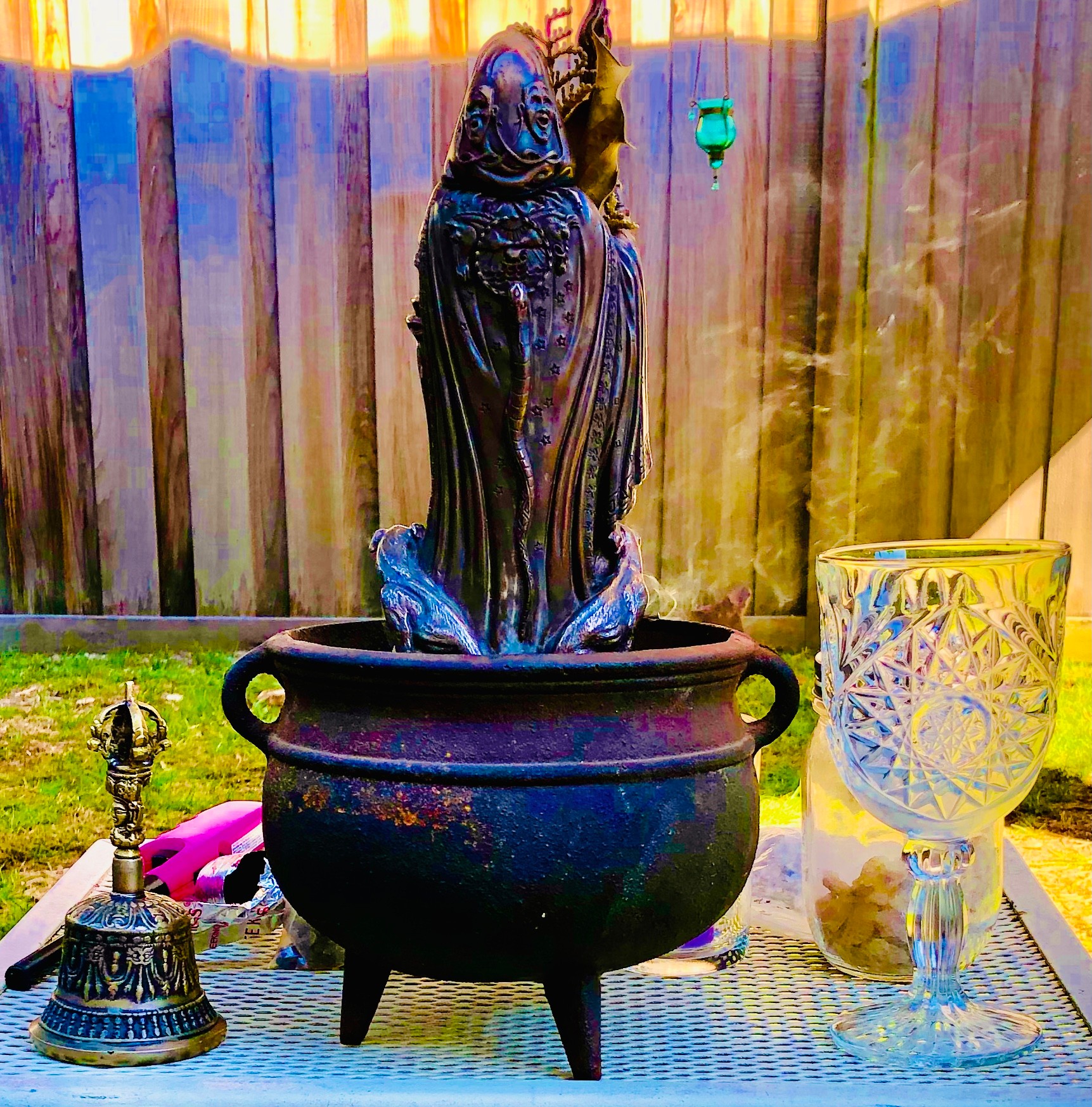 Statue of a figure with a headdress in a large pot, next to a decorative glass and a small bell, outdoors.