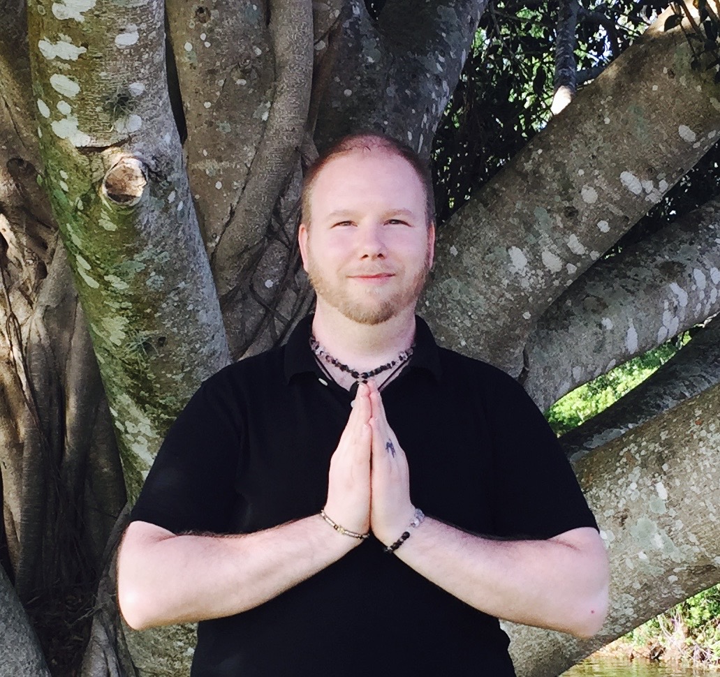 Person with hands in prayer position standing outdoors near a large tree with multiple branches.