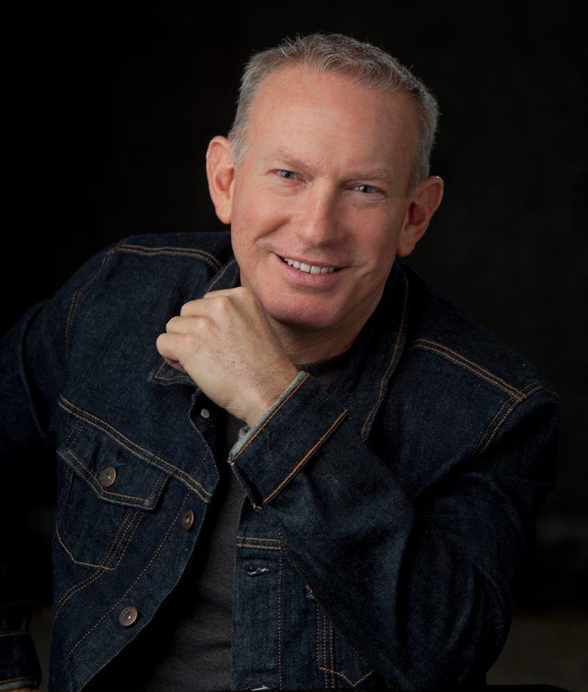 Man with short gray hair smiling, wearing a dark denim jacket, resting his chin on his hand, against a dark background.