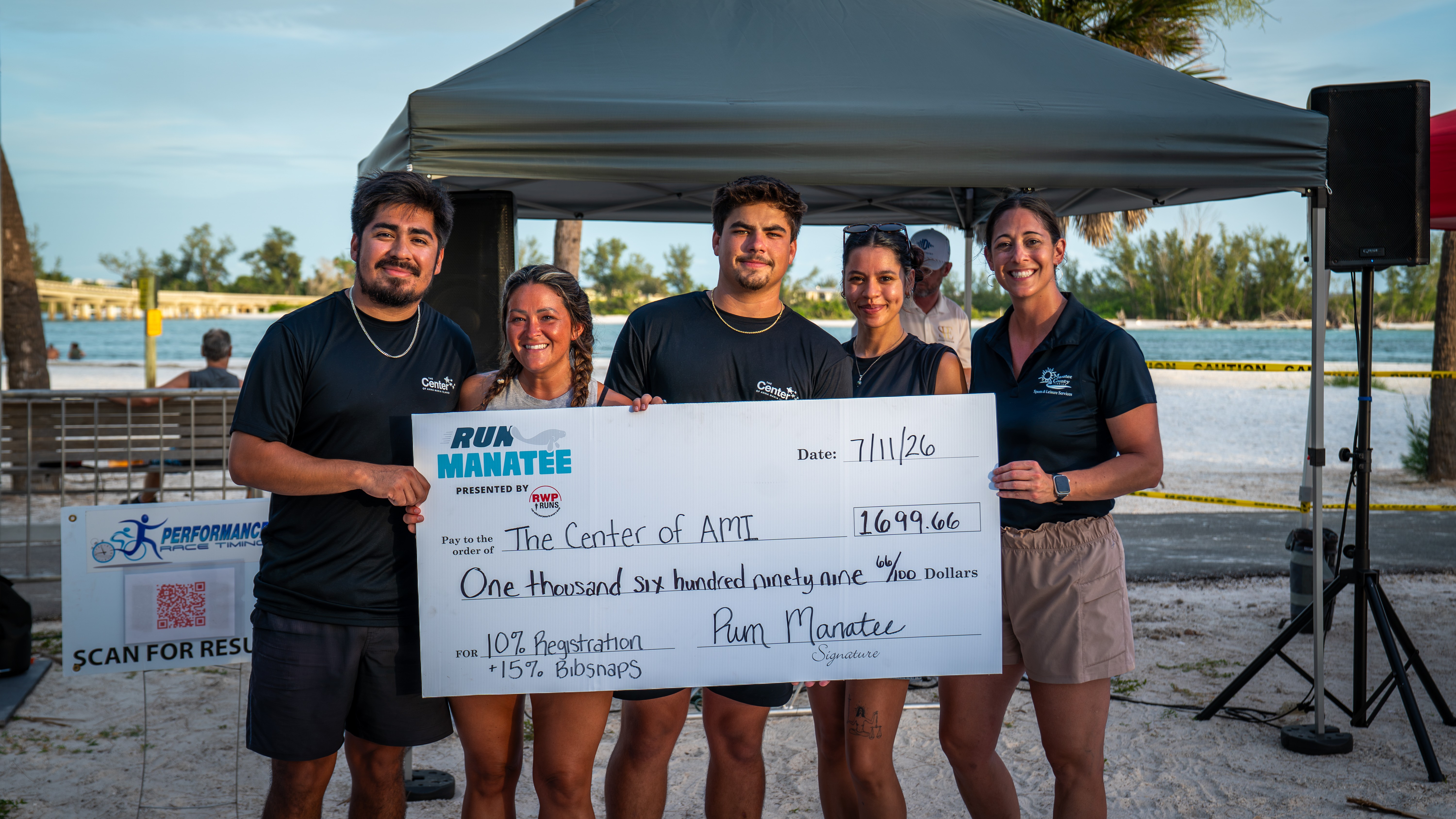 Group of five people holding a large check outdoors near a body of water, with a tent and trees in the background.