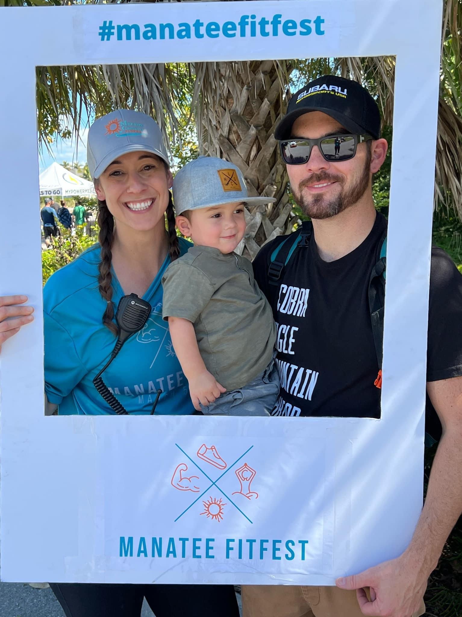 Family of three smiling behind a frame with #manateefitfest and MANATEE FITFEST written on it, outdoors with trees and blue sky.