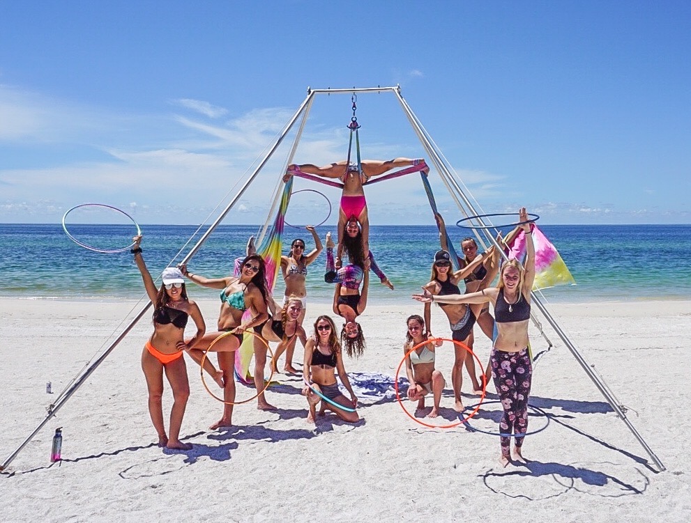 Group of people performing acrobatics on a beach with a metal frame, ocean, and blue sky in background.
