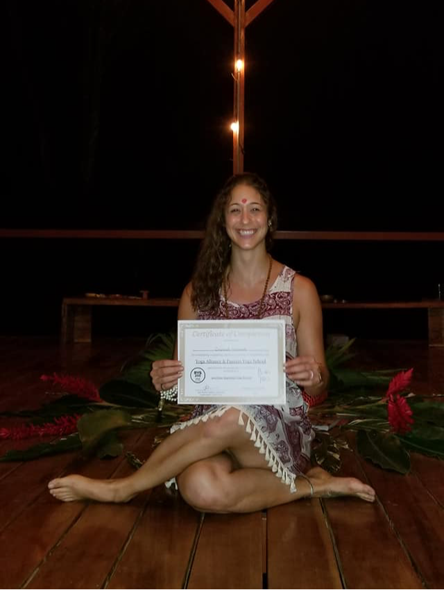 Young woman sitting cross-legged on a wooden floor, holding a certificate, smiling, with plants and a dark background.