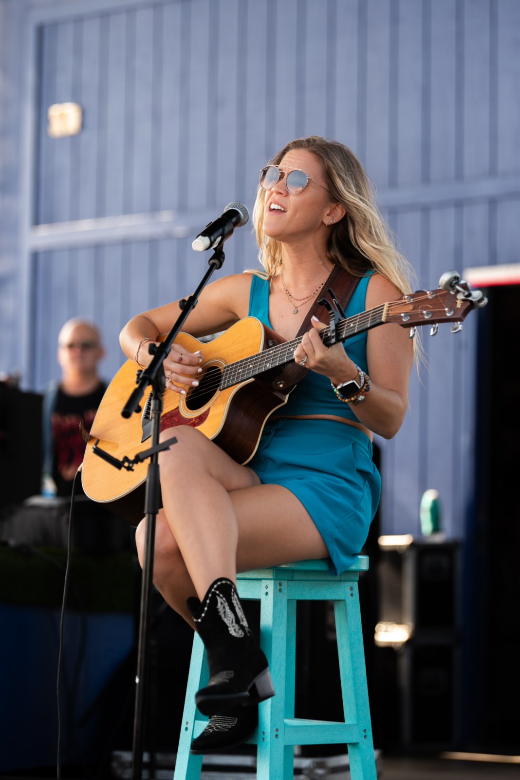 Woman playing guitar and singing into microphone outdoors, wearing sunglasses and a blue dress, sitting on a stool.