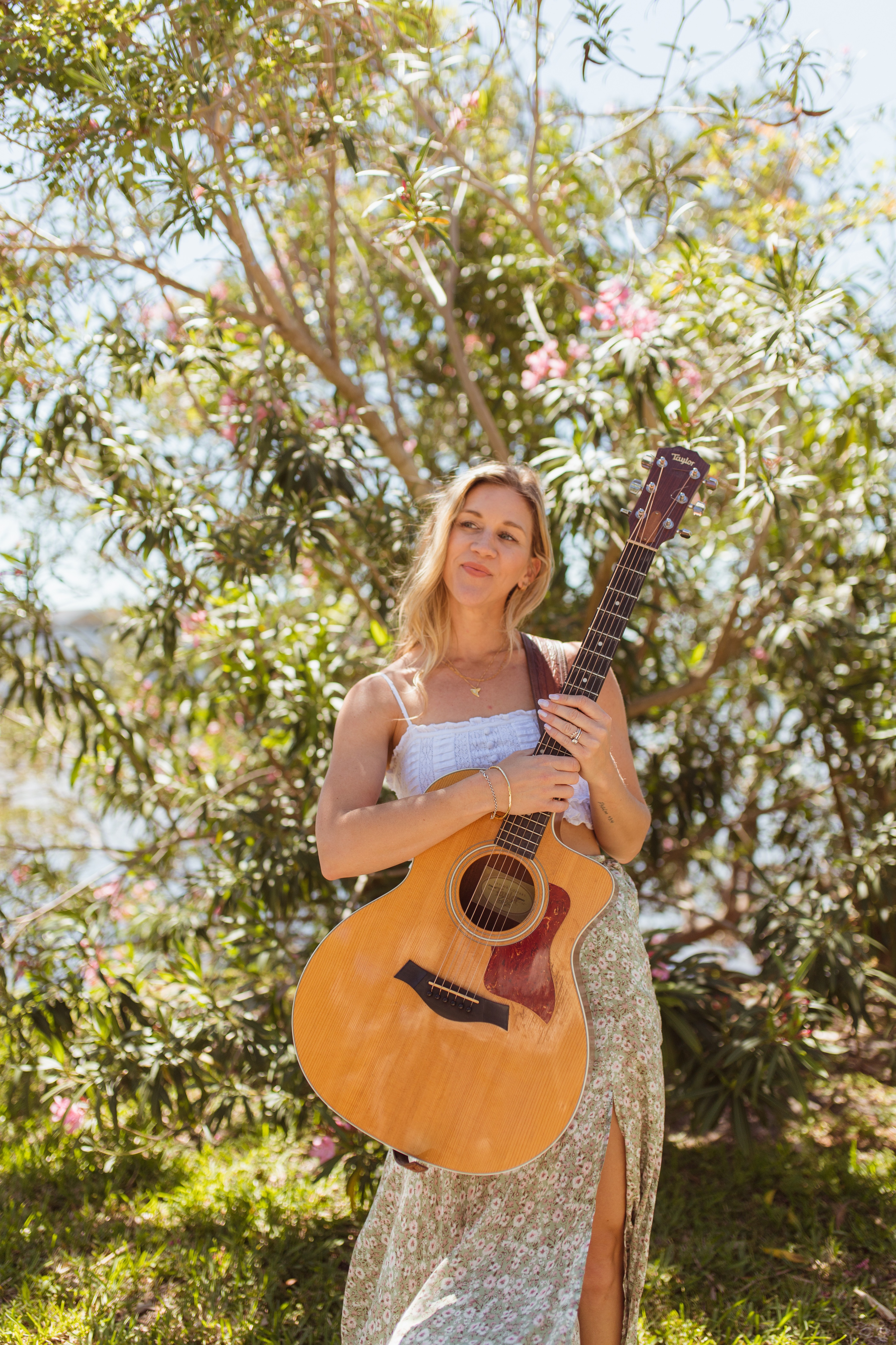 Young woman holding an acoustic guitar outdoors with trees and pink flowers in background.