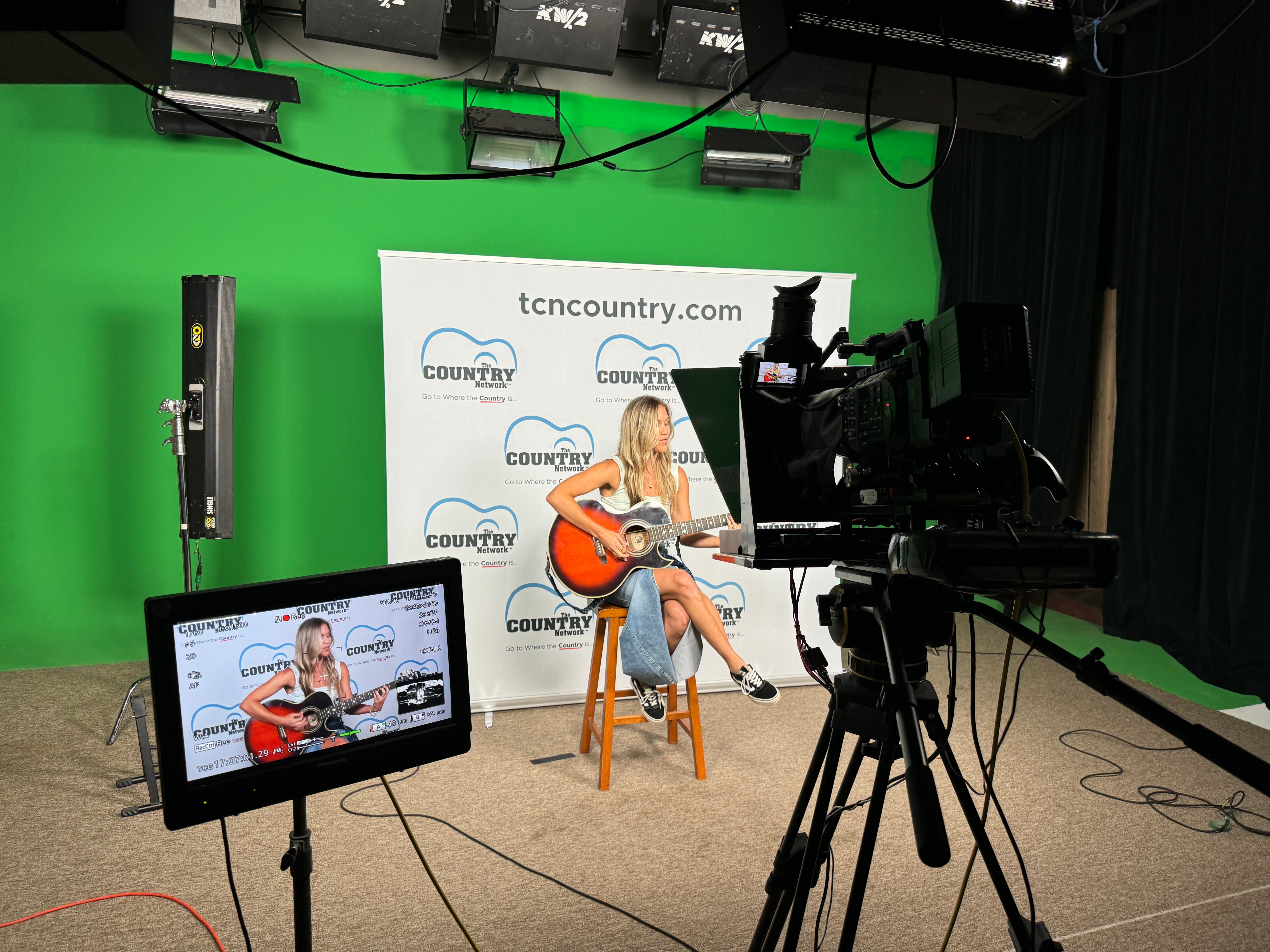 Person playing guitar on a stool in front of a green screen with a backdrop, camera recording the scene.