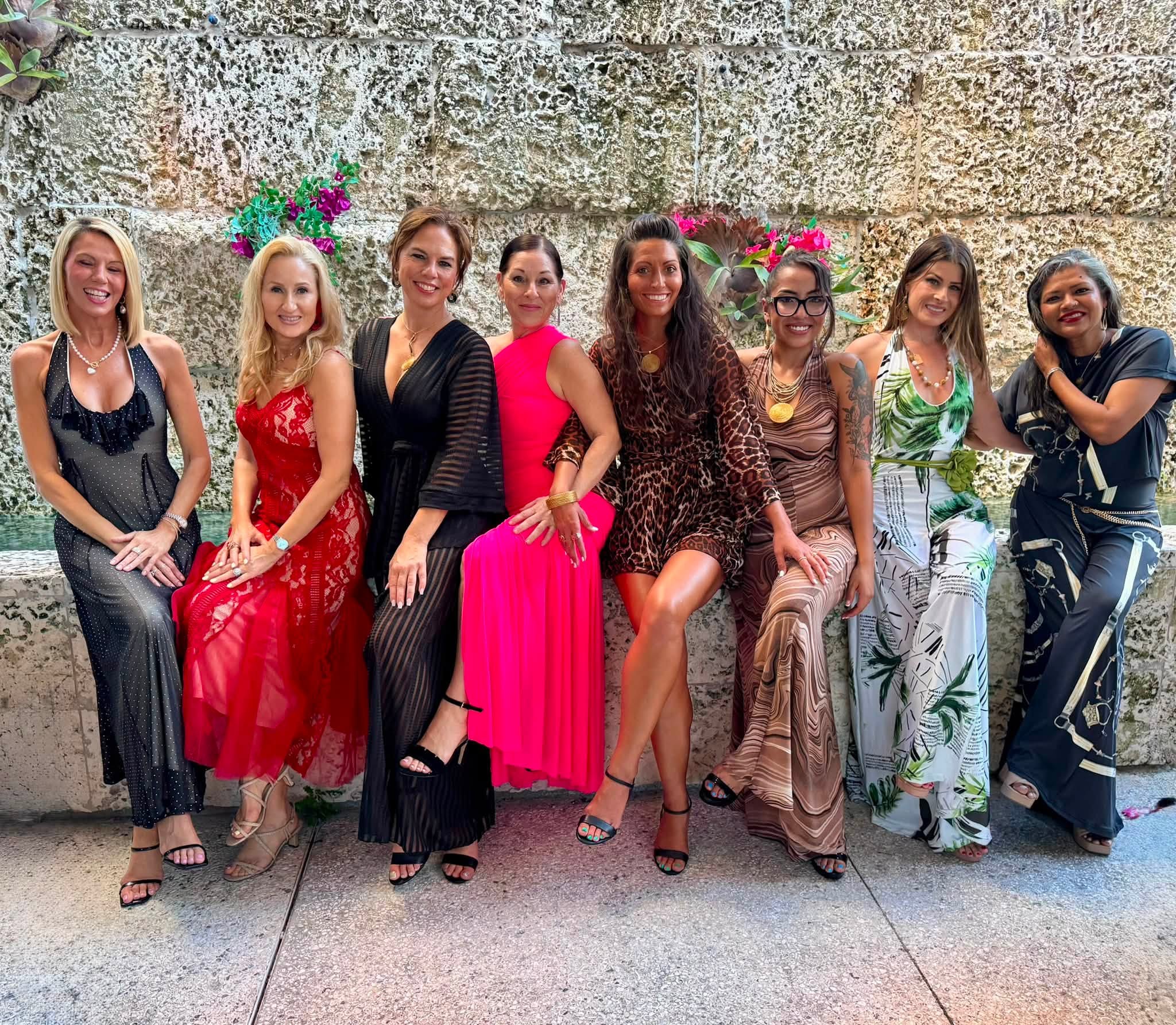 Eight women standing outdoors against a stone wall, dressed in colorful outfits, smiling, with flowers in the background.