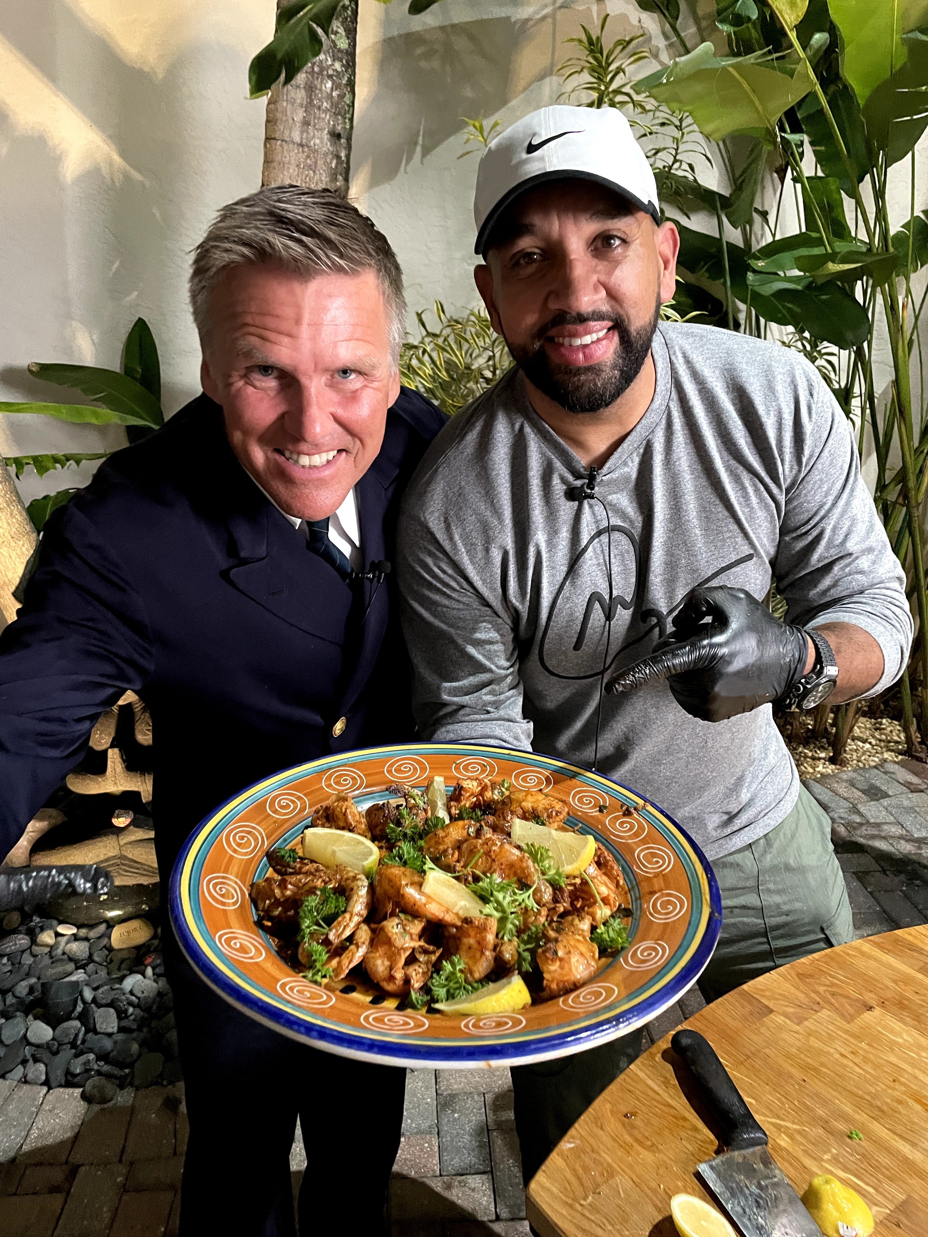 Two men smiling behind a plate of food outdoors, surrounded by plants, with a wooden table in front.