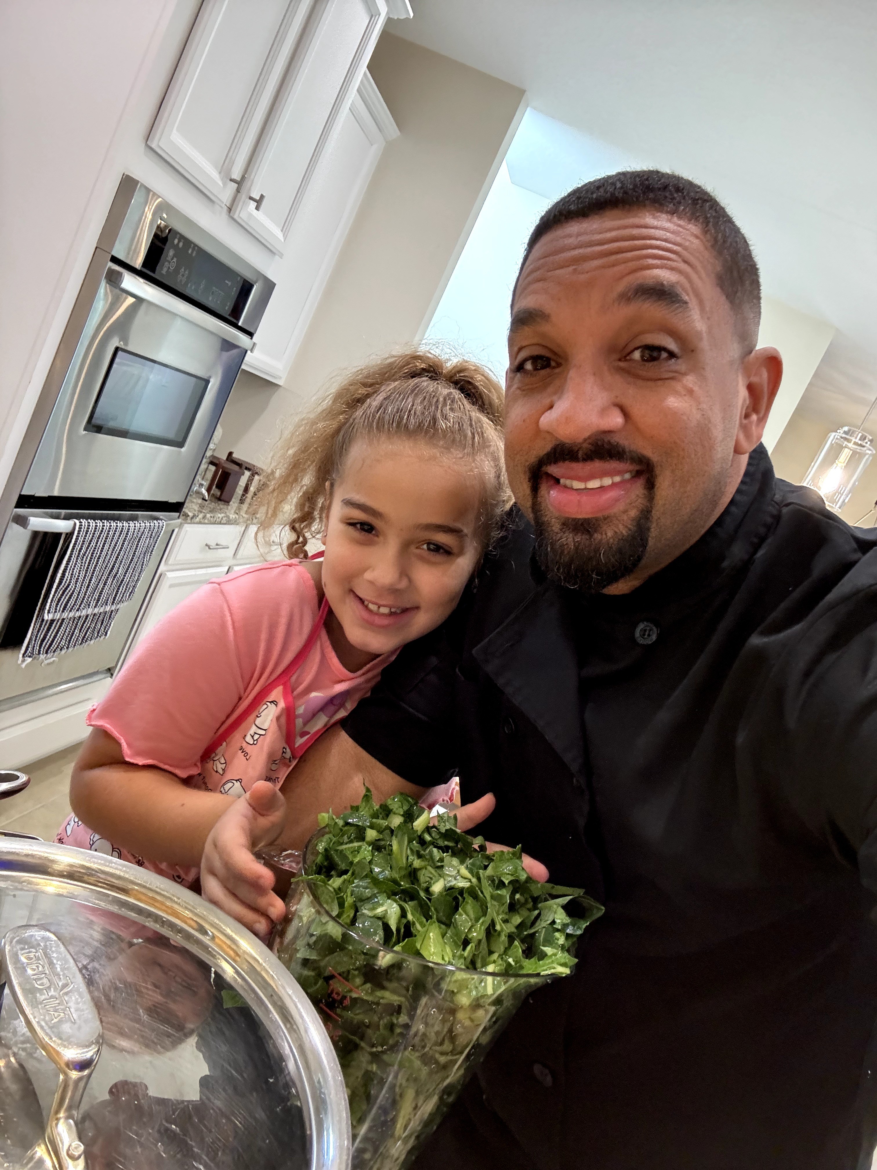 A man and a young girl smiling in a kitchen, holding a bunch of leafy greens, with kitchen appliances in the background.