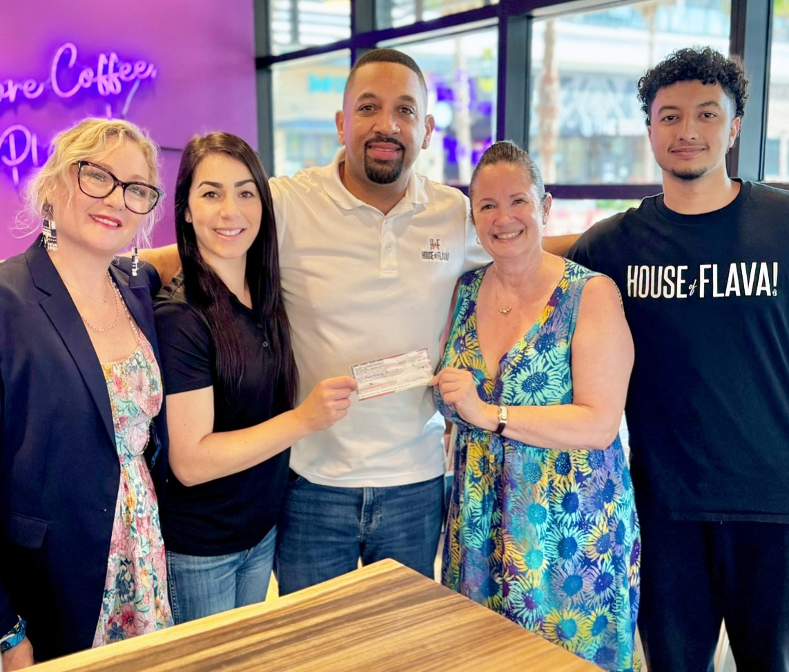Group of five people smiling, holding a check, inside a cafe with neon sign, colorful window, and wooden table.