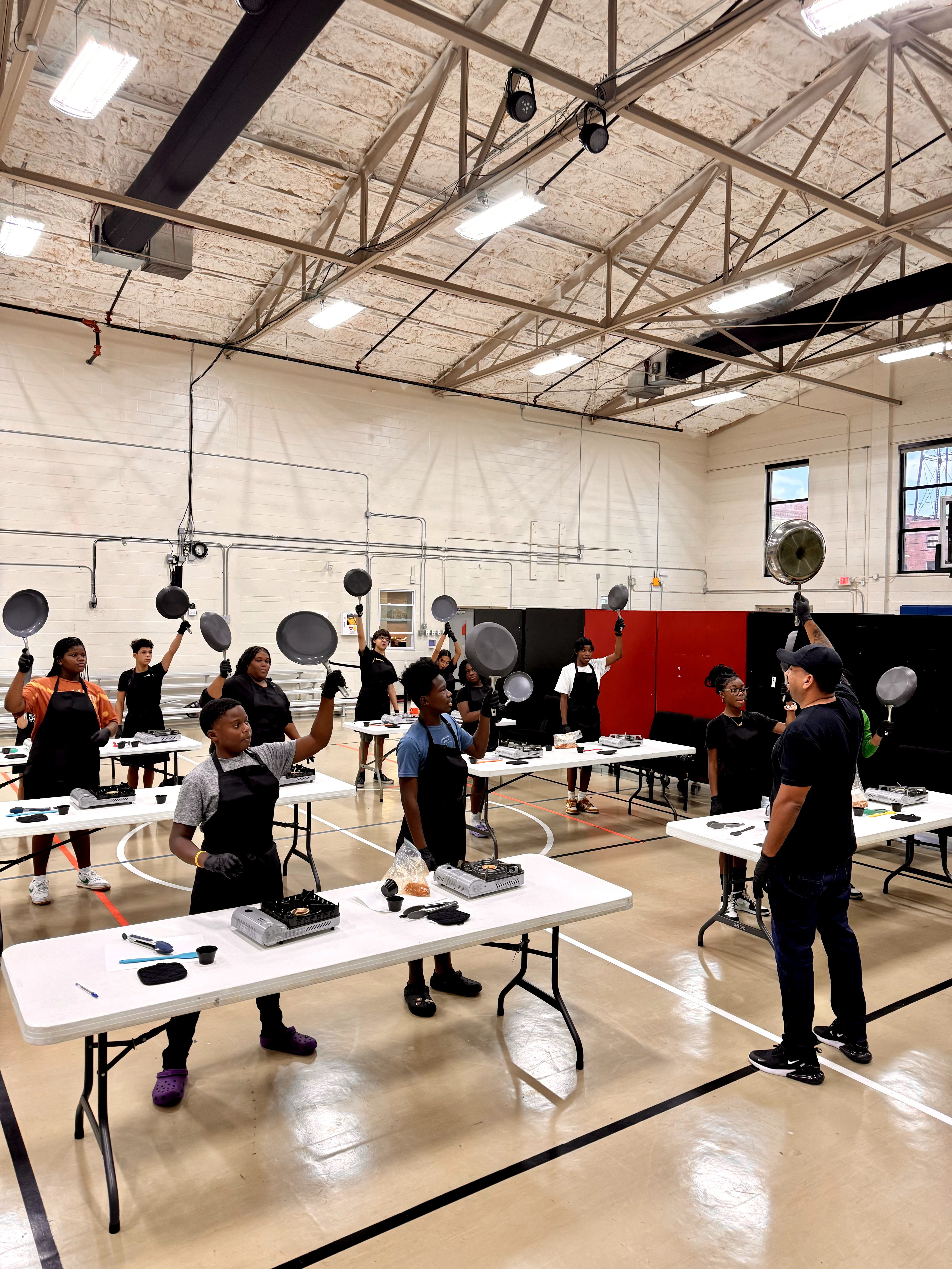 People in a gymnasium participating in a fitness class with black and white equipment, high ceiling, and large windows.