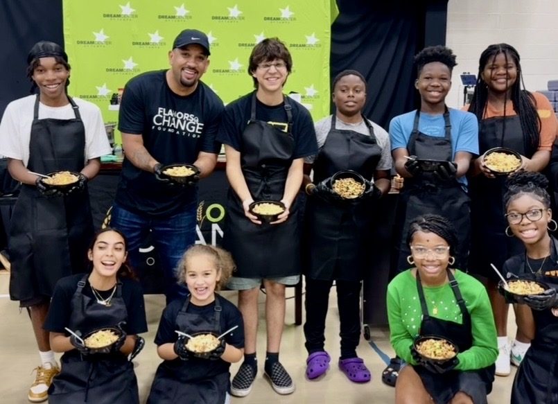 Group of children and two adults holding bowls of food, standing in front of a green backdrop with stars.