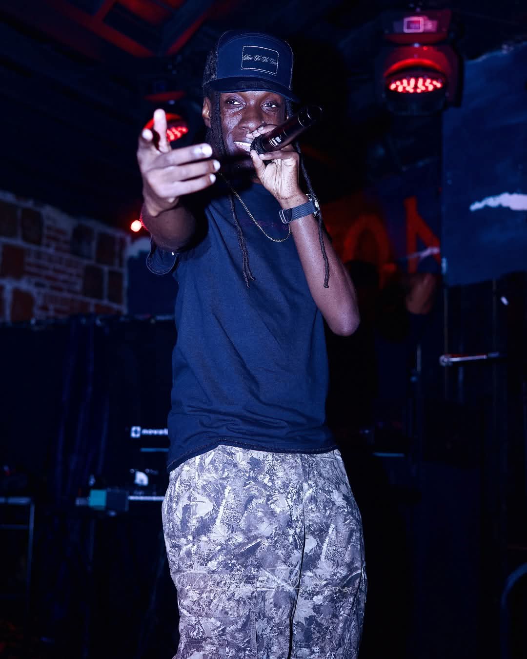 Person wearing a black cap and dark shirt, holding a microphone and pointing forward in a dimly lit setting.