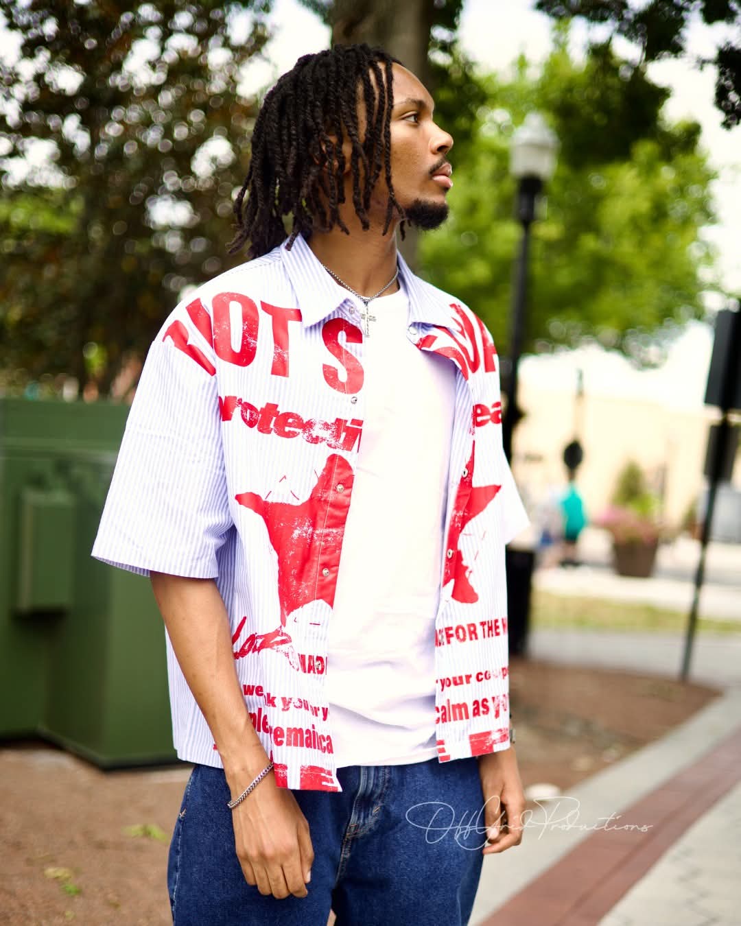 Man with dreadlocks wearing a white shirt with red text and graphics, standing outdoors with trees and a lamp post in background.