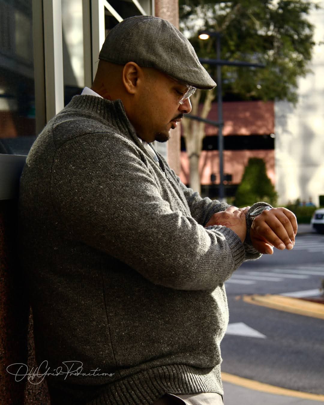Man wearing a gray sweater and flat cap looking at his watch outdoors, with trees and buildings in background.