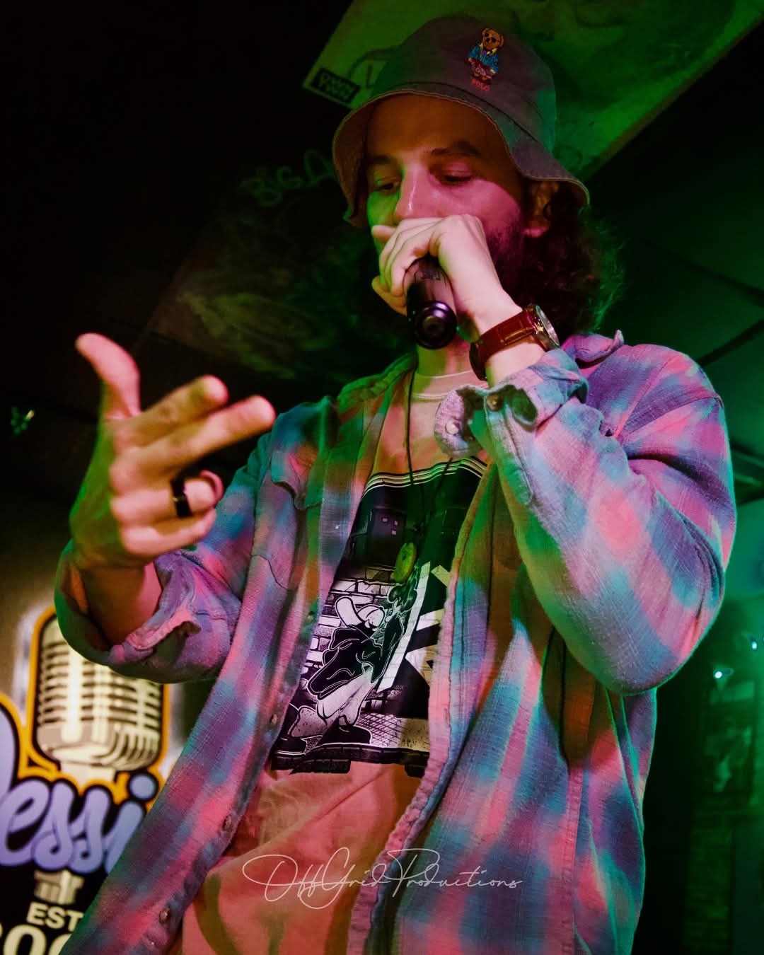 Person wearing a hat and plaid shirt, holding a microphone and performing on stage with green lighting.
