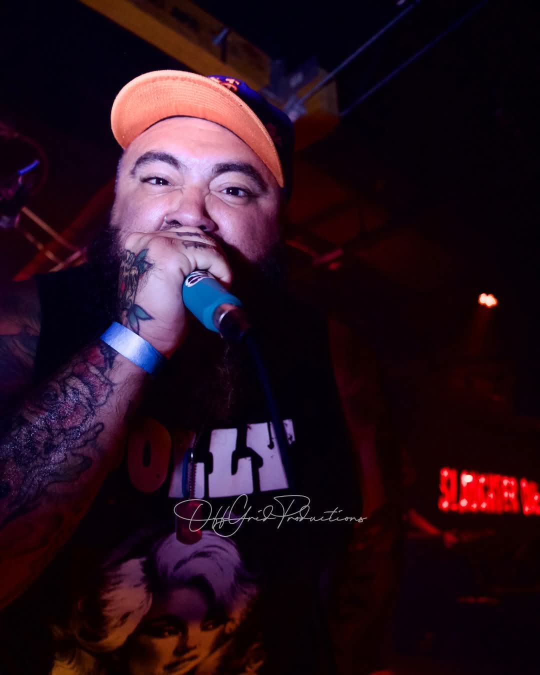 Person singing into a microphone, wearing a cap, with tattoos visible on arm, in a dimly lit setting.