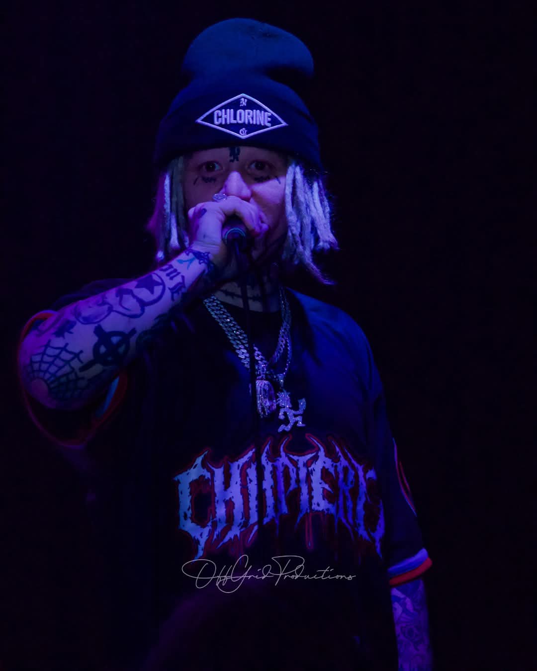 Person with tattoos, wearing a beanie and chain, holding a finger to their lips, dark background, purple lighting.