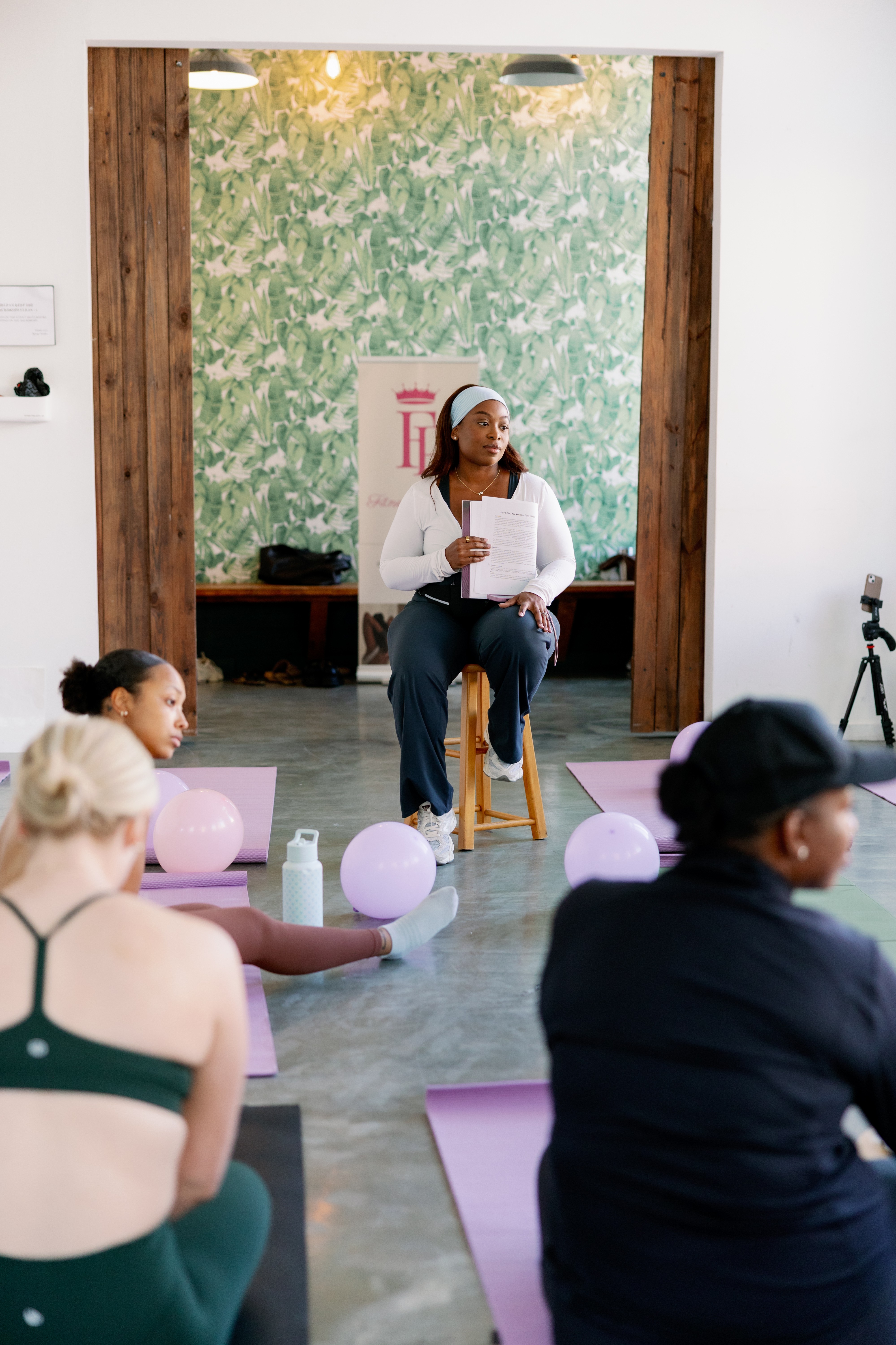 Woman sitting on a stool in front of a green wall, holding a book, with people seated on yoga mats in foreground.