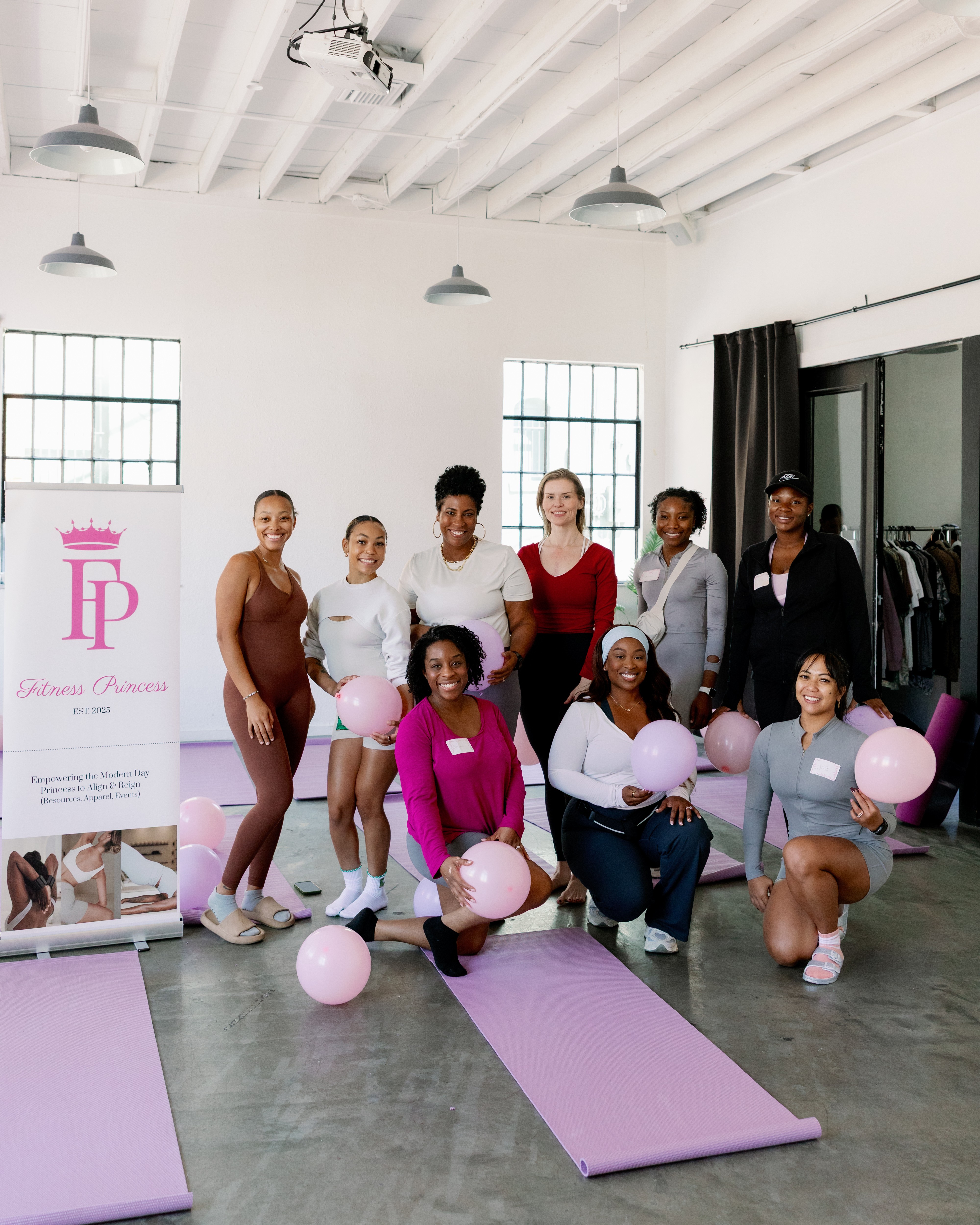 Group of eight women in workout clothes holding pink and white balloons in a bright studio with yoga mats and a banner.
