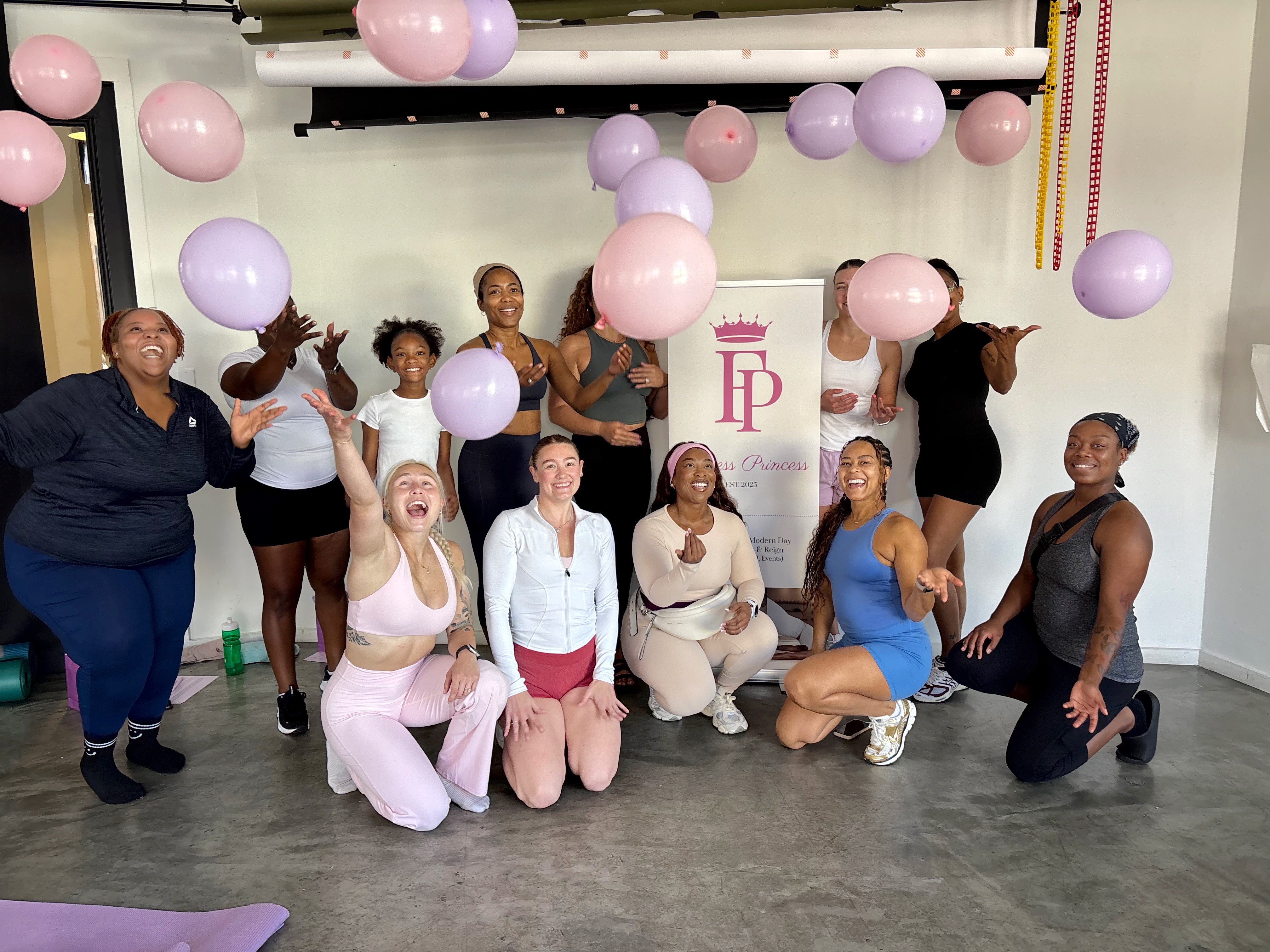 Group of nine women in a fitness studio with pink and purple balloons, some standing and some kneeling, smiling and celebrating.