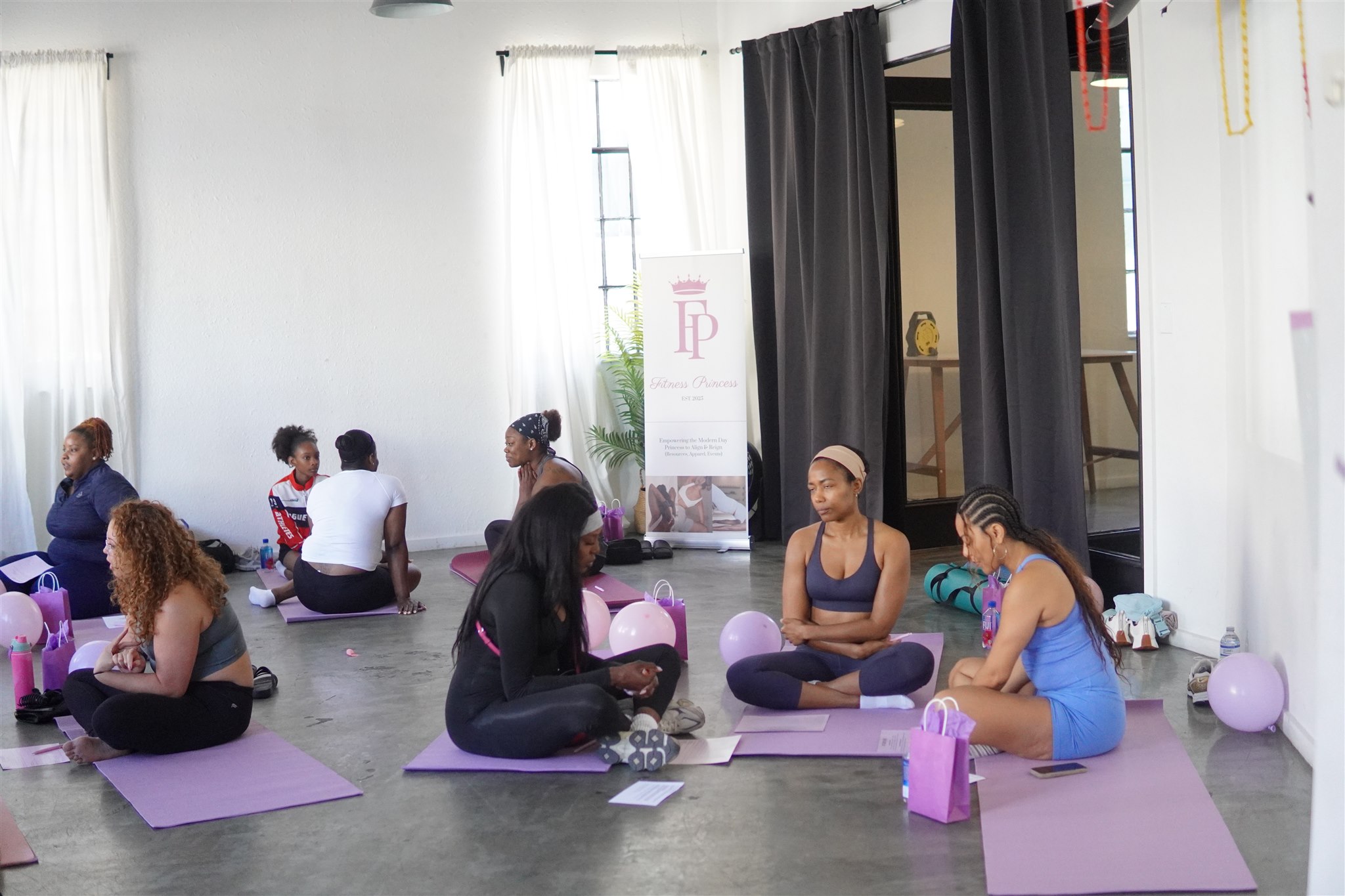 People participating in a yoga class in a bright studio with large windows, sitting on purple mats, some with balloons nearby.