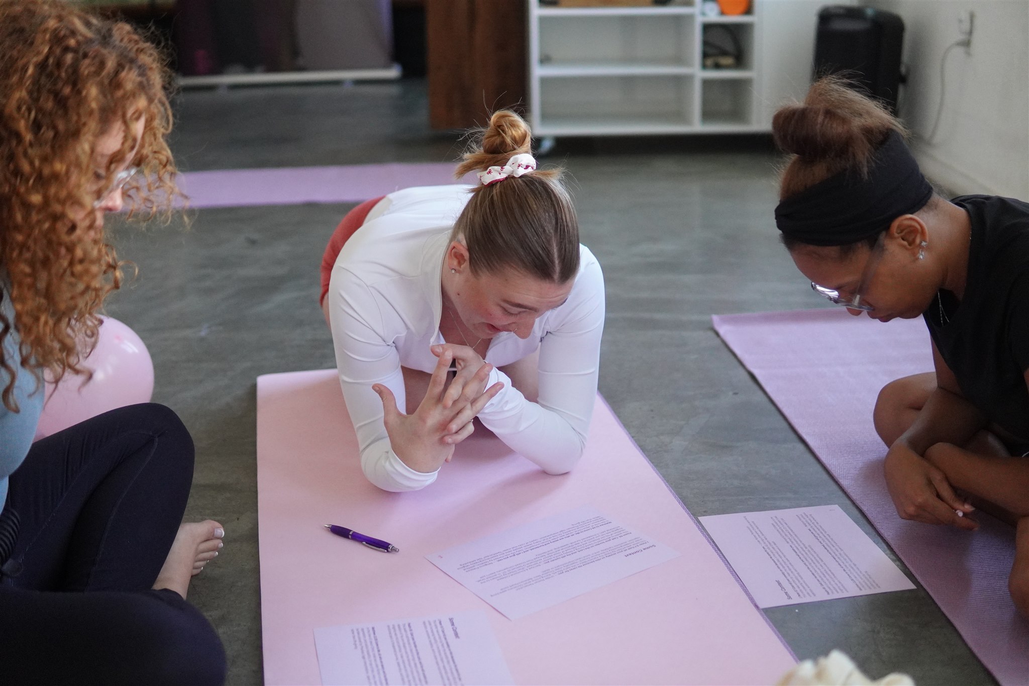 Three women sitting on the floor, one in white with hair in a bun, two in dark clothing, on yoga mats, with papers and a pen nearby