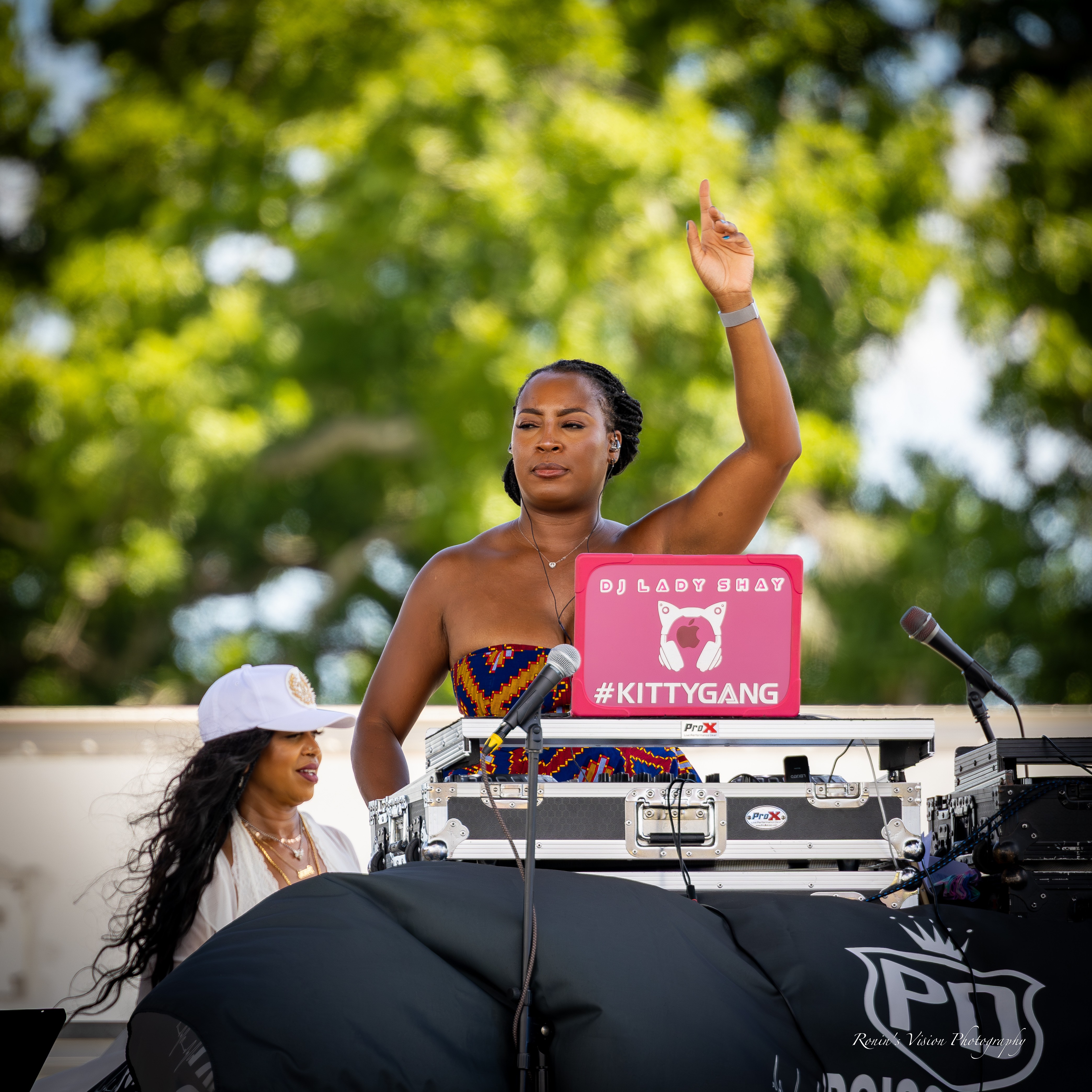 Female DJ with a laptop and equipment outdoors, raising her right hand, with a woman in a white hat nearby, trees in background.