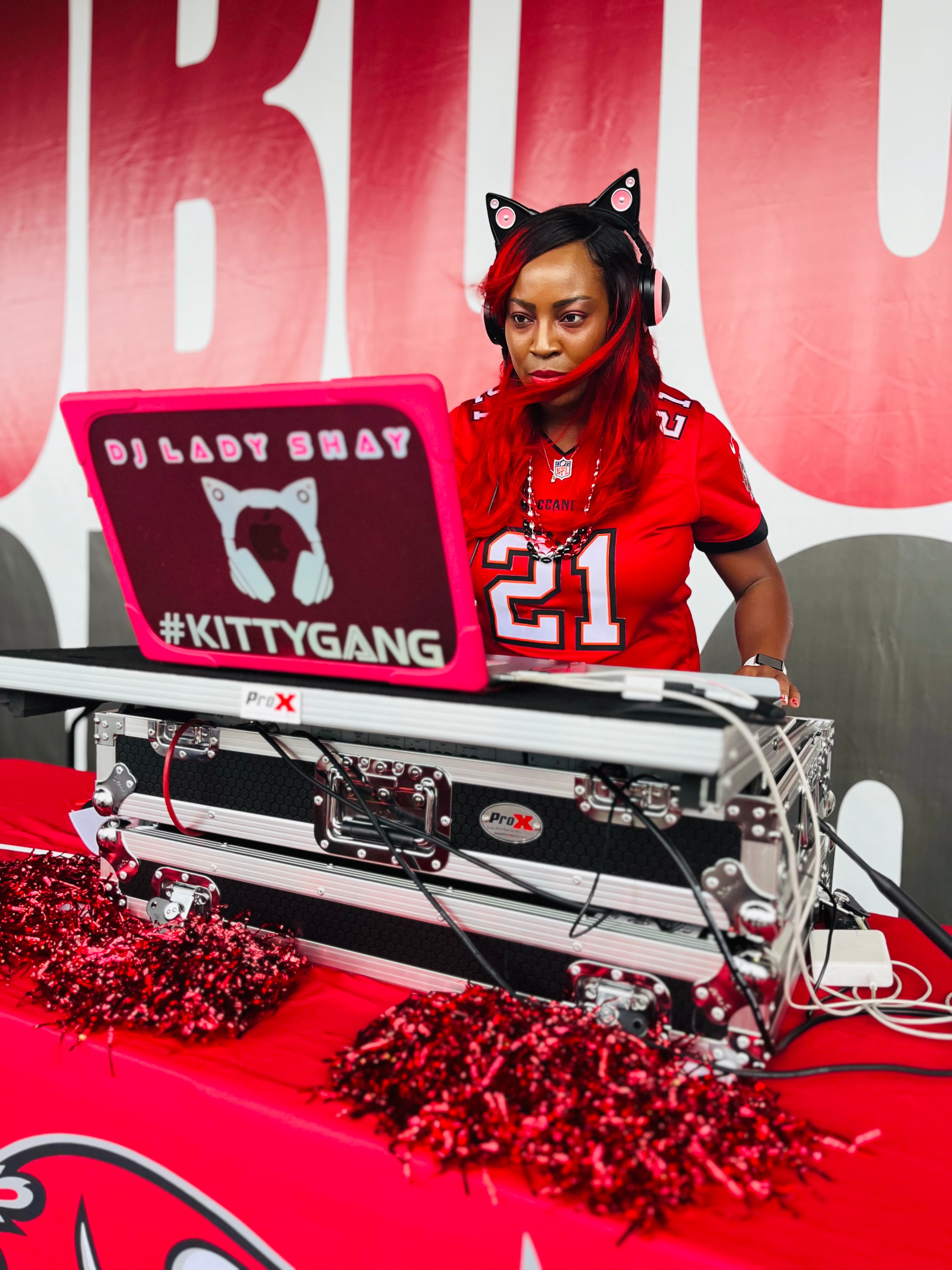Woman with red hair and cat ear headband DJing at a table with red decorations, wearing a red sports jersey, in front of a red and white background.