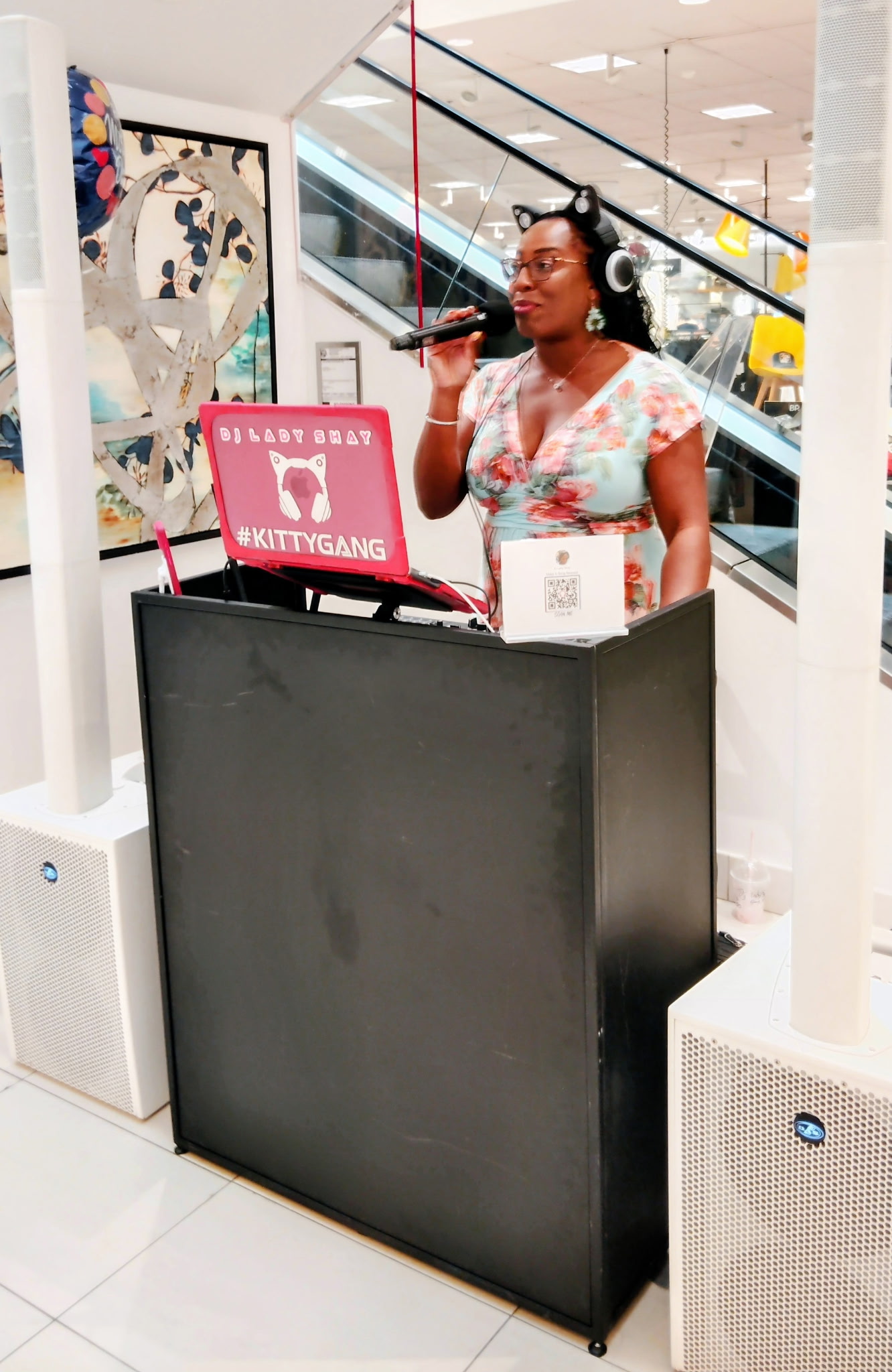 Woman with headphones singing into microphone at a karaoke station in a shopping mall, with a pink sign and artwork in background.