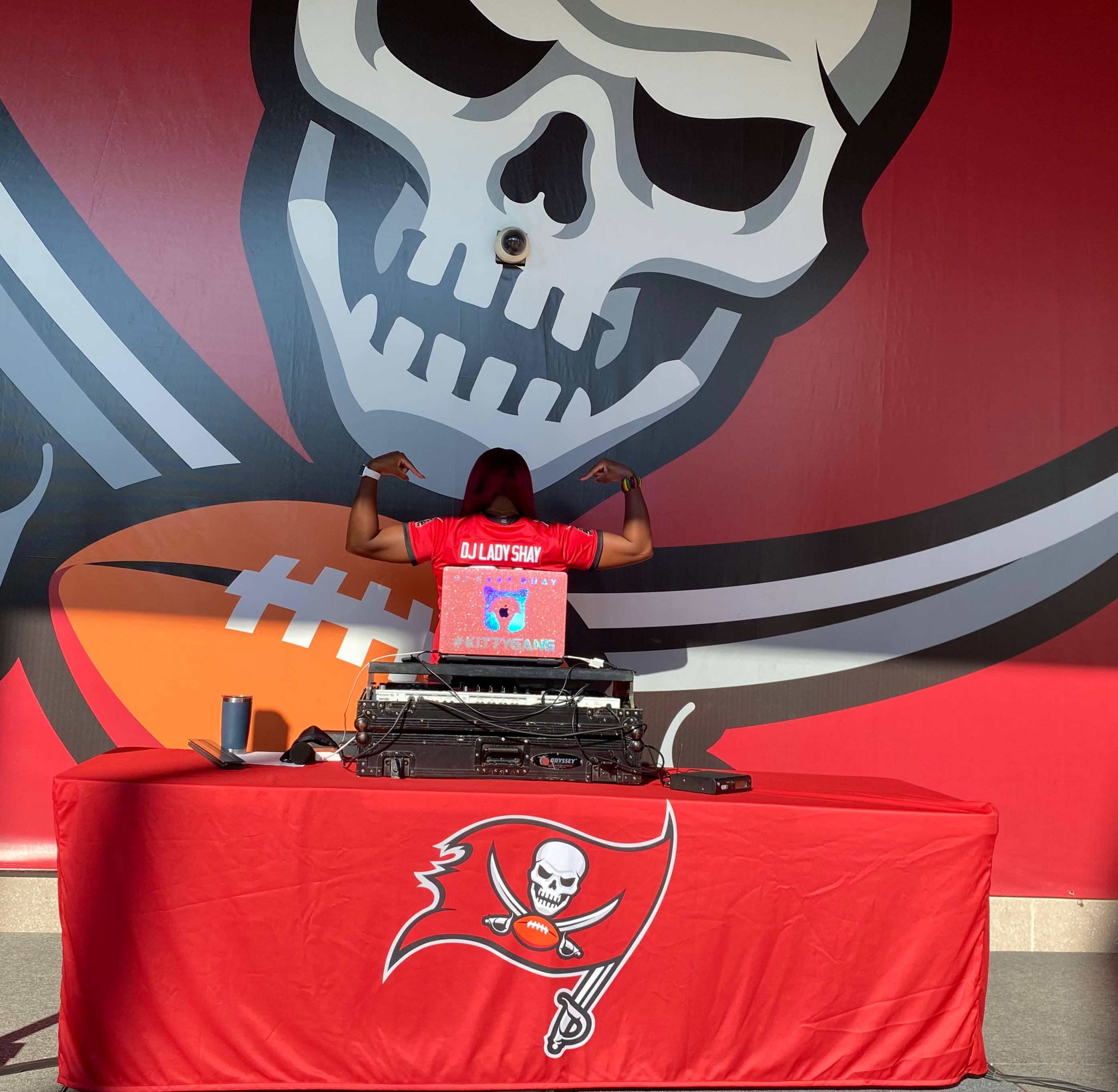 Person with arms raised standing behind a DJ setup on a red table, with a large skull and bat logo on a wall behind them.
