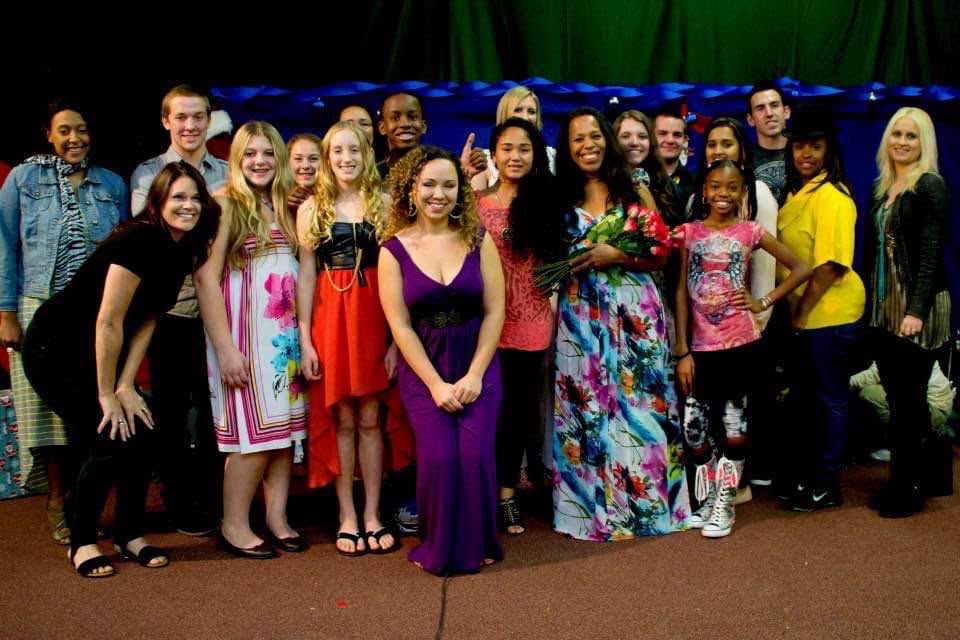 Group of diverse people standing together on stage, smiling, with colorful background and green curtain.