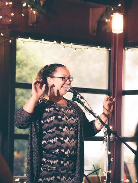 Woman with glasses smiling and raising her hand, standing in front of a microphone indoors with windows and warm lighting.