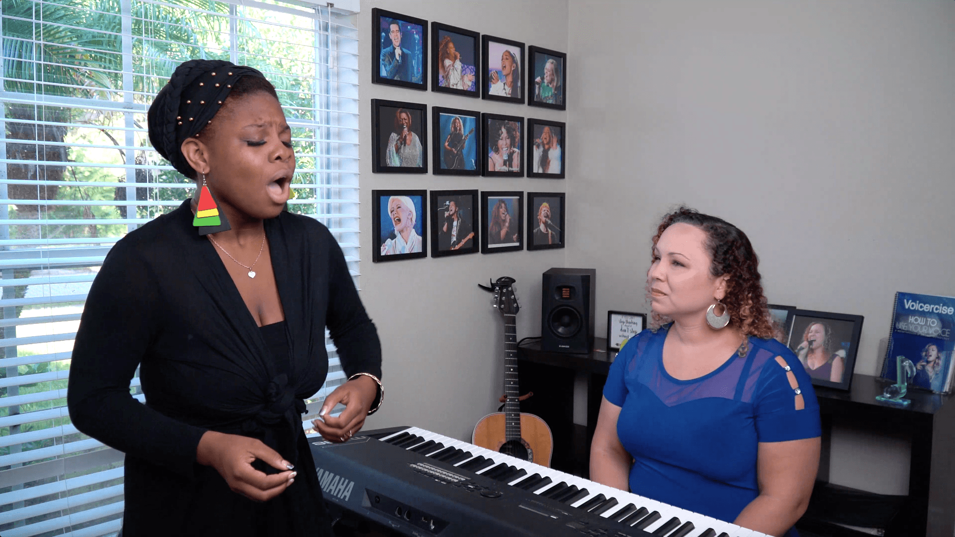 Two women, one singing and the other playing keyboard, in a room with framed photos on the wall.