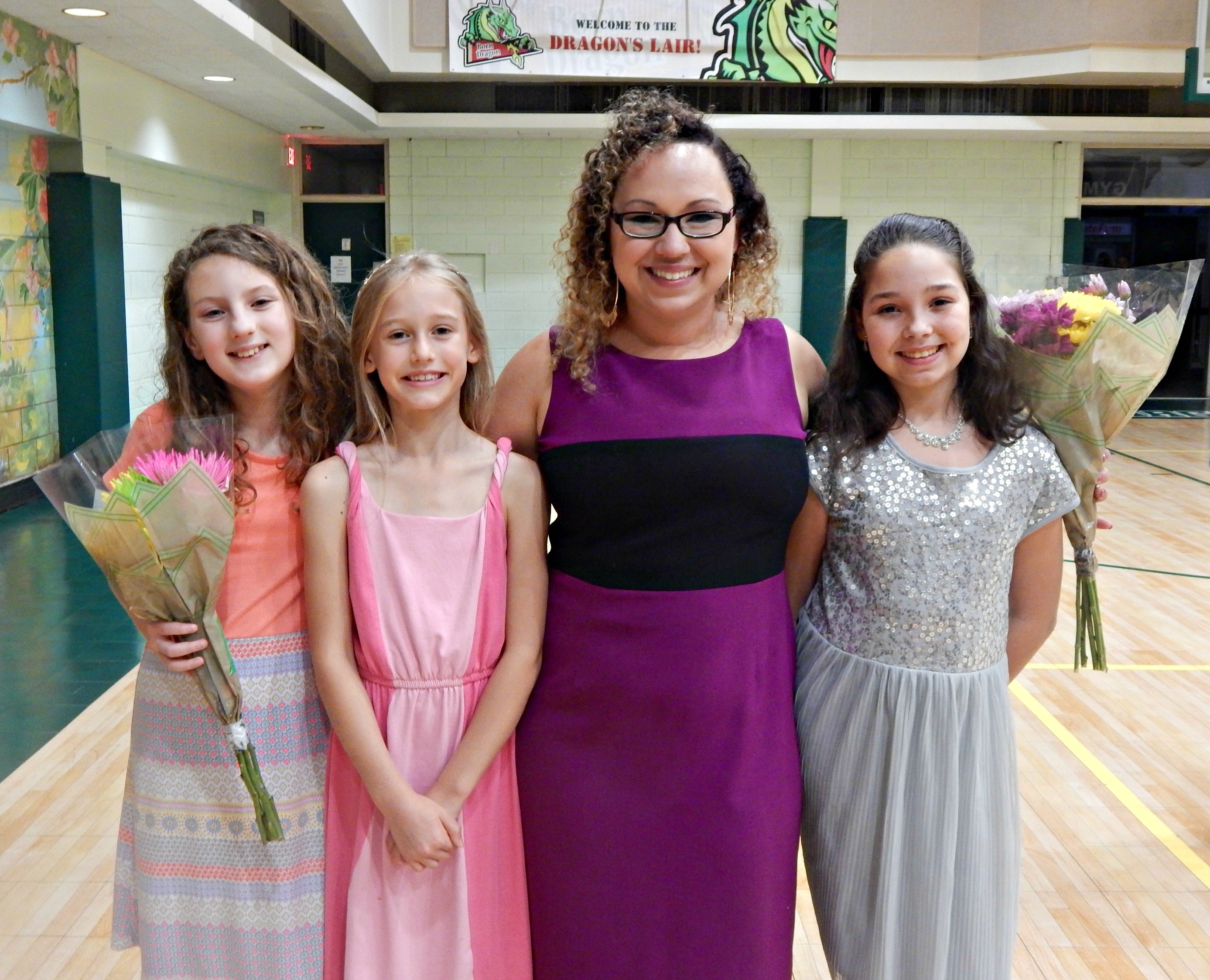 Four people, three young girls and one adult woman, standing indoors, smiling, with two holding bouquets of flowers.